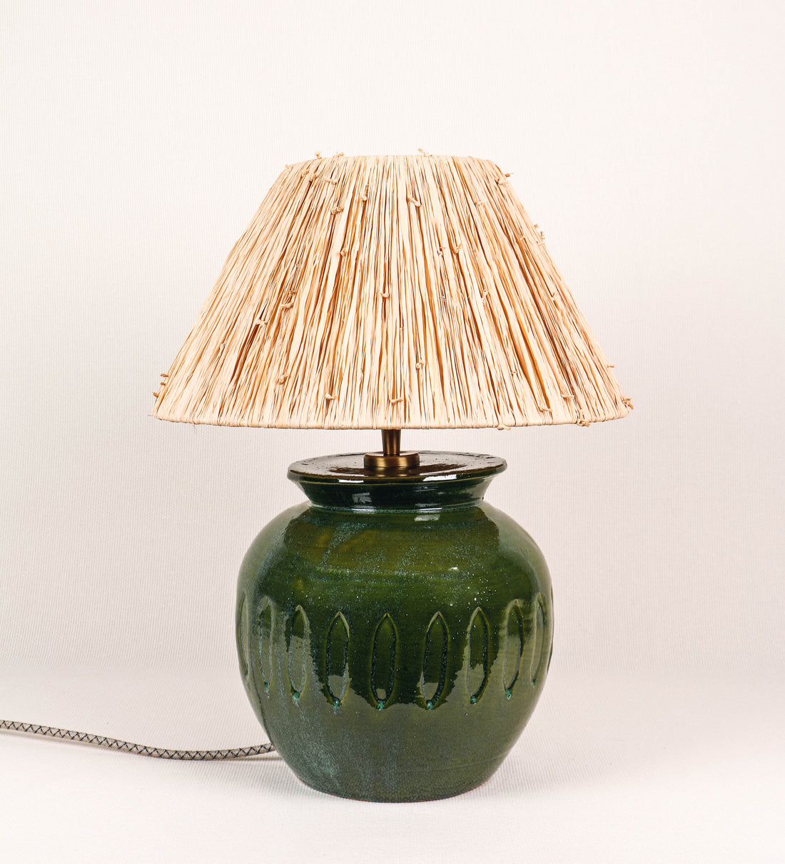 Candeeiro de mesa com um design rústico e sofisticado. A base do candeeiro é de barro vidrado de tom verde vidro. A forma da base é arredondada com uma borda superior mais estreita, lembrando um vaso tradicional. A superfície da base é decorada com um padrão de relevos ovais. O suporte da lâmpada é em latão escovado e é manipulável. O cabo é revestido têxtil com um padrão trançado de linhas castanhas e beges, com interruptor e ficha transparentes. O abatjour é cónico, de ráfia com nós.