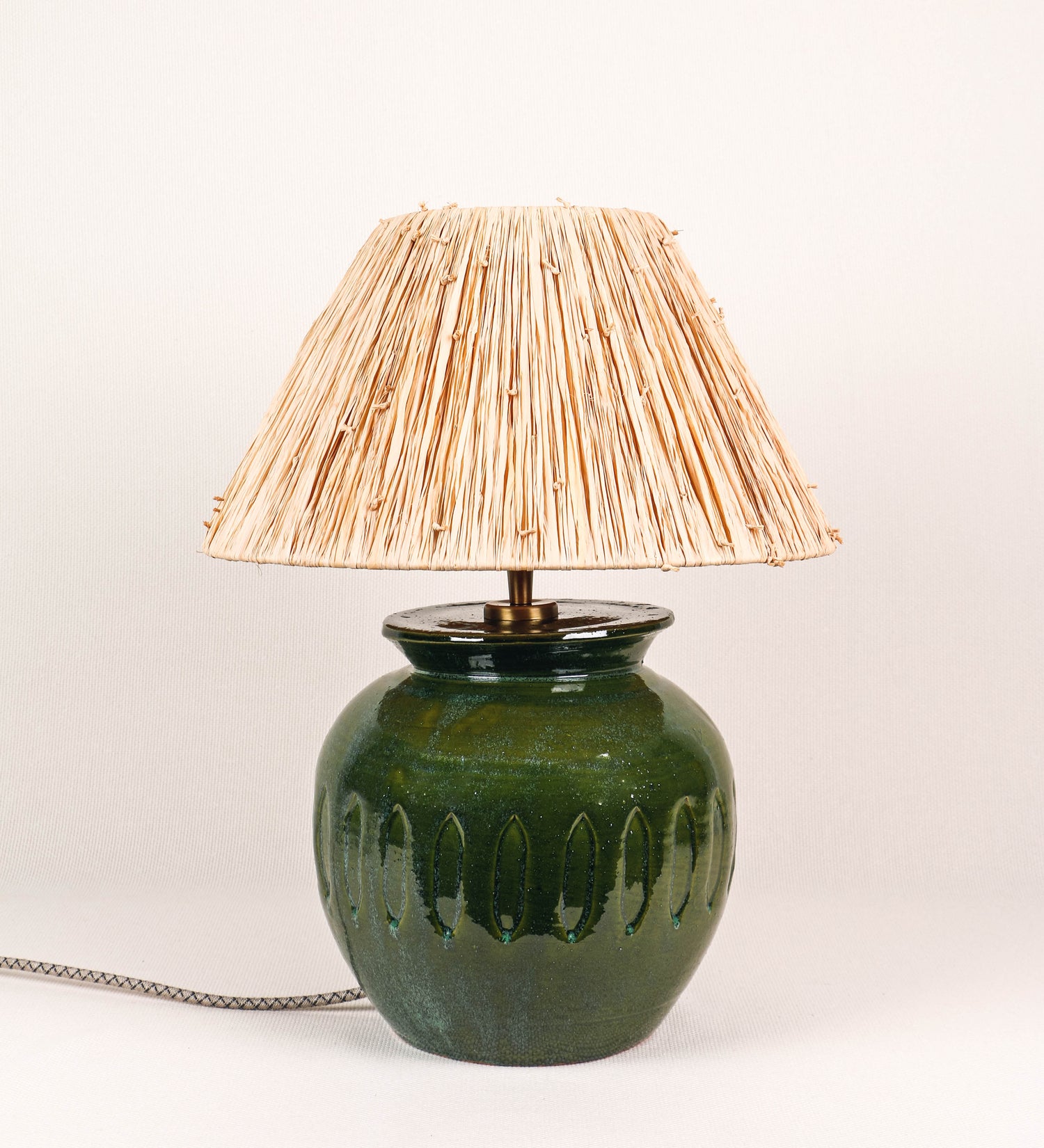 Candeeiro de mesa com um design rústico e sofisticado. A base do candeeiro é de barro vidrado de tom verde vidro. A forma da base é arredondada com uma borda superior mais estreita, lembrando um vaso tradicional. A superfície da base é decorada com um padrão de relevos ovais. O suporte da lâmpada é em latão escovado e é manipulável. O cabo é revestido têxtil com um padrão trançado de linhas castanhas e beges, com interruptor e ficha transparentes. O abatjour é cónico, de ráfia com nós.