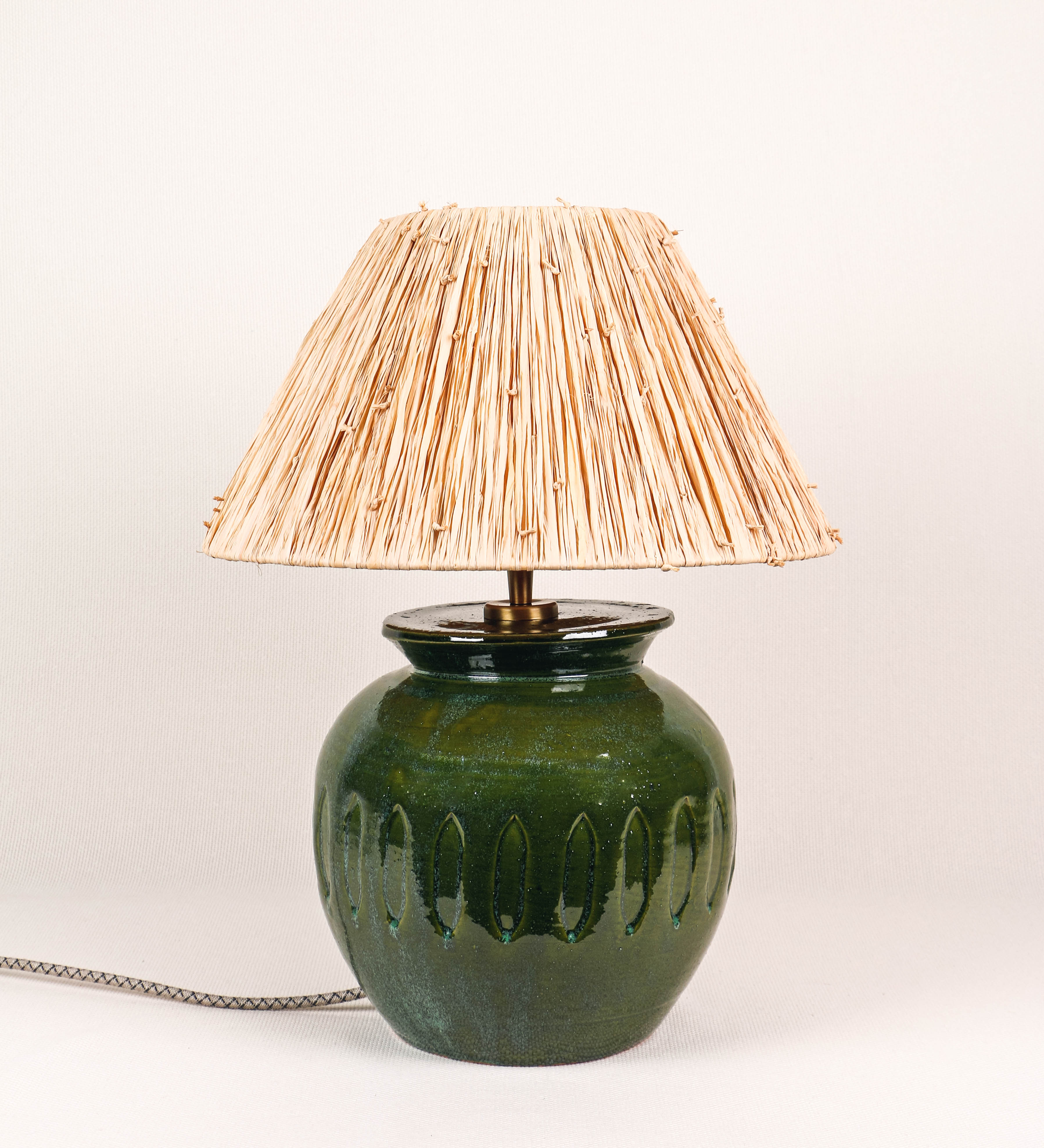 Candeeiro de mesa com um design rústico e sofisticado. A base do candeeiro é de barro vidrado de tom verde vidro. A forma da base é arredondada com uma borda superior mais estreita, lembrando um vaso tradicional. A superfície da base é decorada com um padrão de relevos ovais. O suporte da lâmpada é em latão escovado e é manipulável. O cabo é revestido têxtil com um padrão trançado de linhas castanhas e beges, com interruptor e ficha transparentes. O abatjour é cónico, de ráfia com nós.