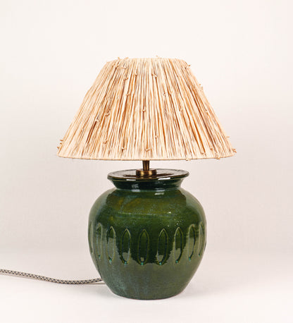 Candeeiro de mesa com um design rústico e sofisticado. A base do candeeiro é de barro vidrado de tom verde vidro. A forma da base é arredondada com uma borda superior mais estreita, lembrando um vaso tradicional. A superfície da base é decorada com um padrão de relevos ovais. O suporte da lâmpada é em latão escovado e é manipulável. O cabo é revestido têxtil com um padrão trançado de linhas castanhas e beges, com interruptor e ficha transparentes. O abatjour é cónico, de ráfia com nós.