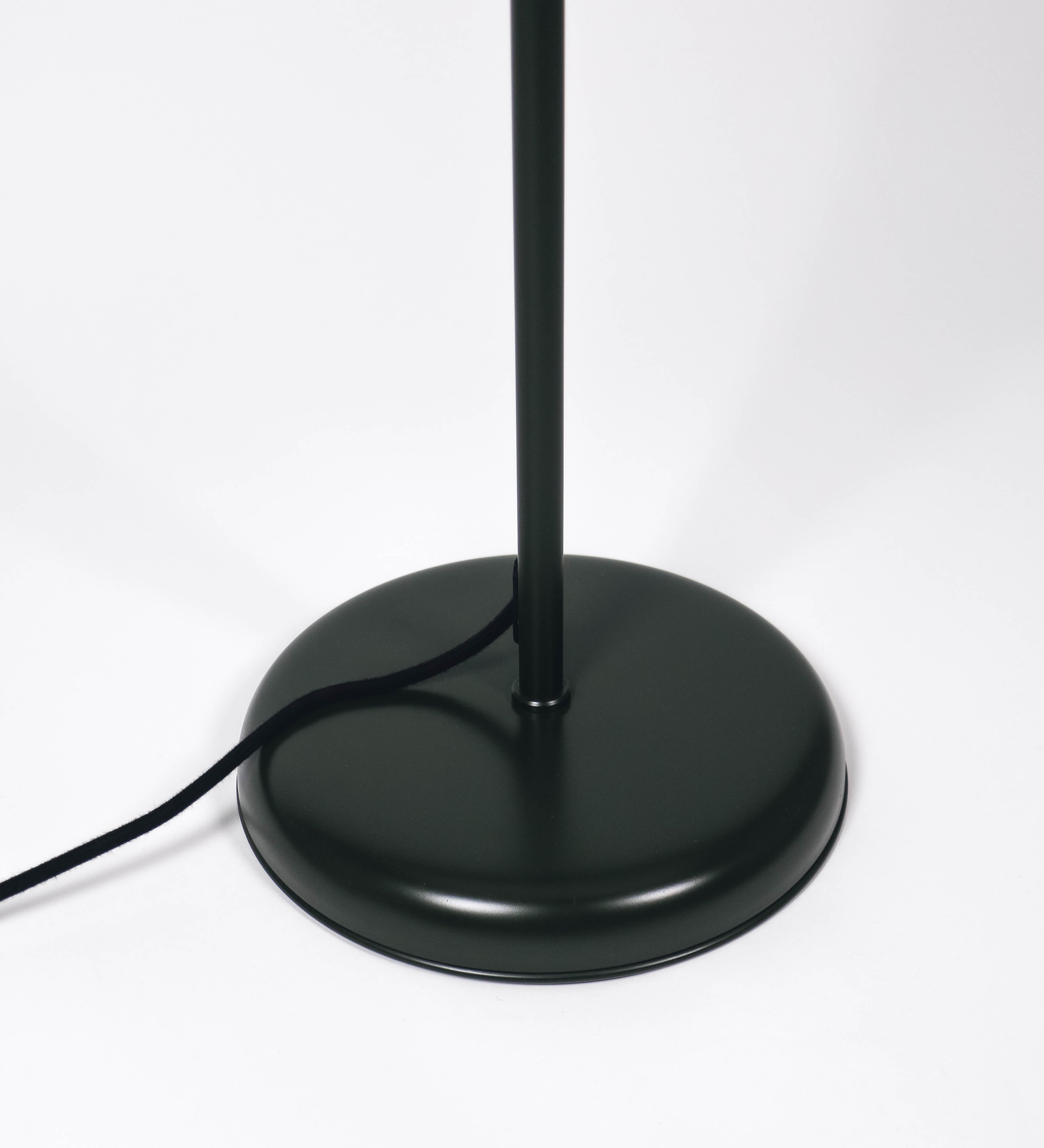Candeeiro de pé com base circular em verde-azeitona. O corpo é fino e vertical, composto por uma haste verde-azeitona, e uma outra haste em níquel escovado, que permite ajustar a altura do candeeiro. A haste ajustável conecta a um braço horizontal, que sustenta o chapéu, e é manipulável. O chapéu é longo e arredondado, em verde-azeitona, e com uma pequena haste com uma esfera na ponta. O interior do chapéu é branco. O cabo é revestido têxtil em preto, com um interruptor de pé preto circular.