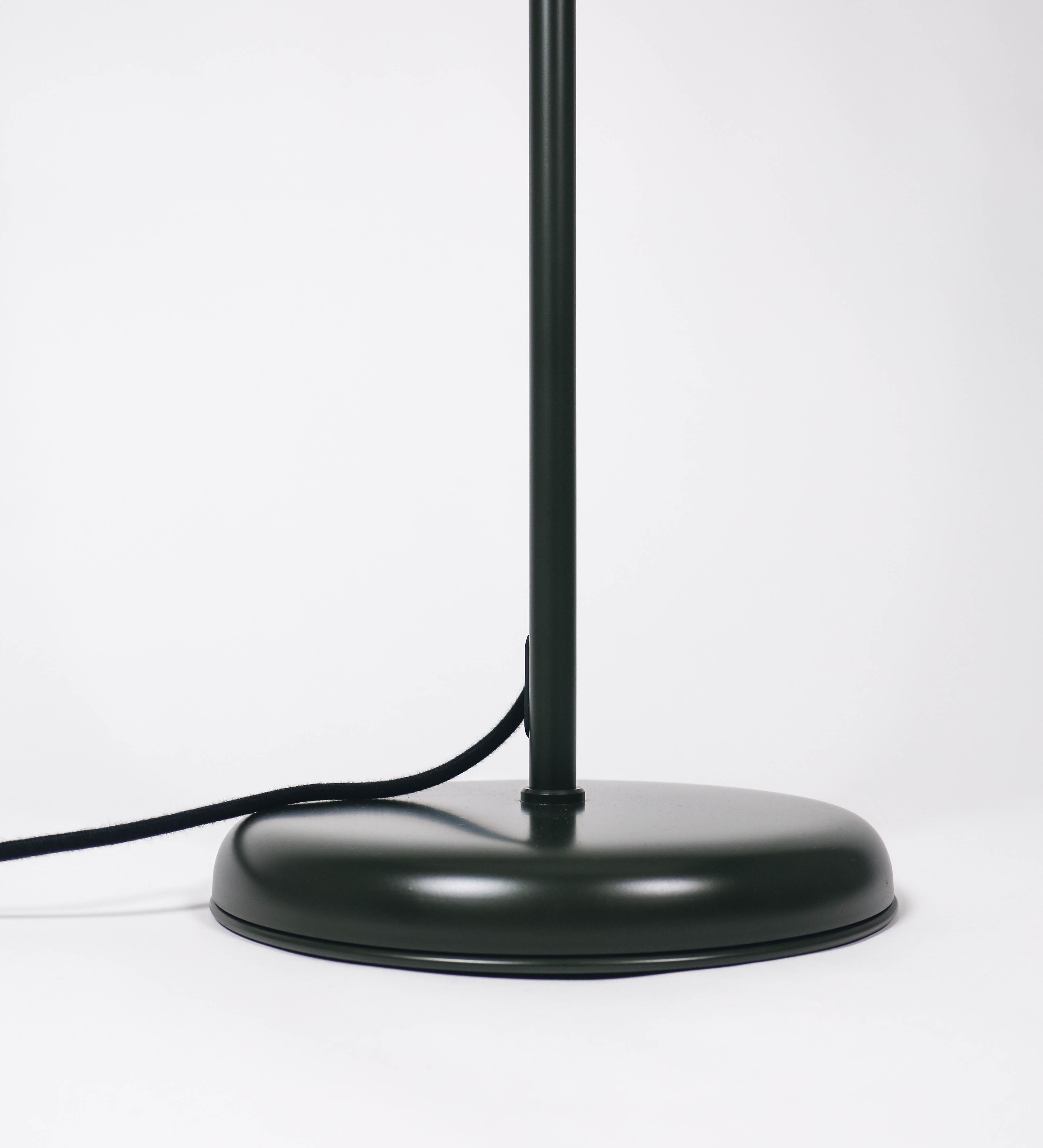Candeeiro de pé com base circular em verde-azeitona. O corpo é fino e vertical, composto por uma haste verde-azeitona, e uma outra haste em níquel escovado, que permite ajustar a altura do candeeiro. A haste ajustável conecta a um braço horizontal, que sustenta o chapéu, e é manipulável. O chapéu é longo e arredondado, em verde-azeitona, e com uma pequena haste com uma esfera na ponta. O interior do chapéu é branco. O cabo é revestido têxtil em preto, com um interruptor de pé preto circular.