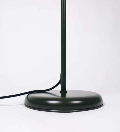Candeeiro de pé com base circular em verde-azeitona. O corpo é fino e vertical, composto por uma haste verde-azeitona, e uma outra haste em níquel escovado, que permite ajustar a altura do candeeiro. A haste ajustável conecta a um braço horizontal, que sustenta o chapéu, e é manipulável. O chapéu é longo e arredondado, em verde-azeitona, e com uma pequena haste com uma esfera na ponta. O interior do chapéu é branco. O cabo é revestido têxtil em preto, com um interruptor de pé preto circular.