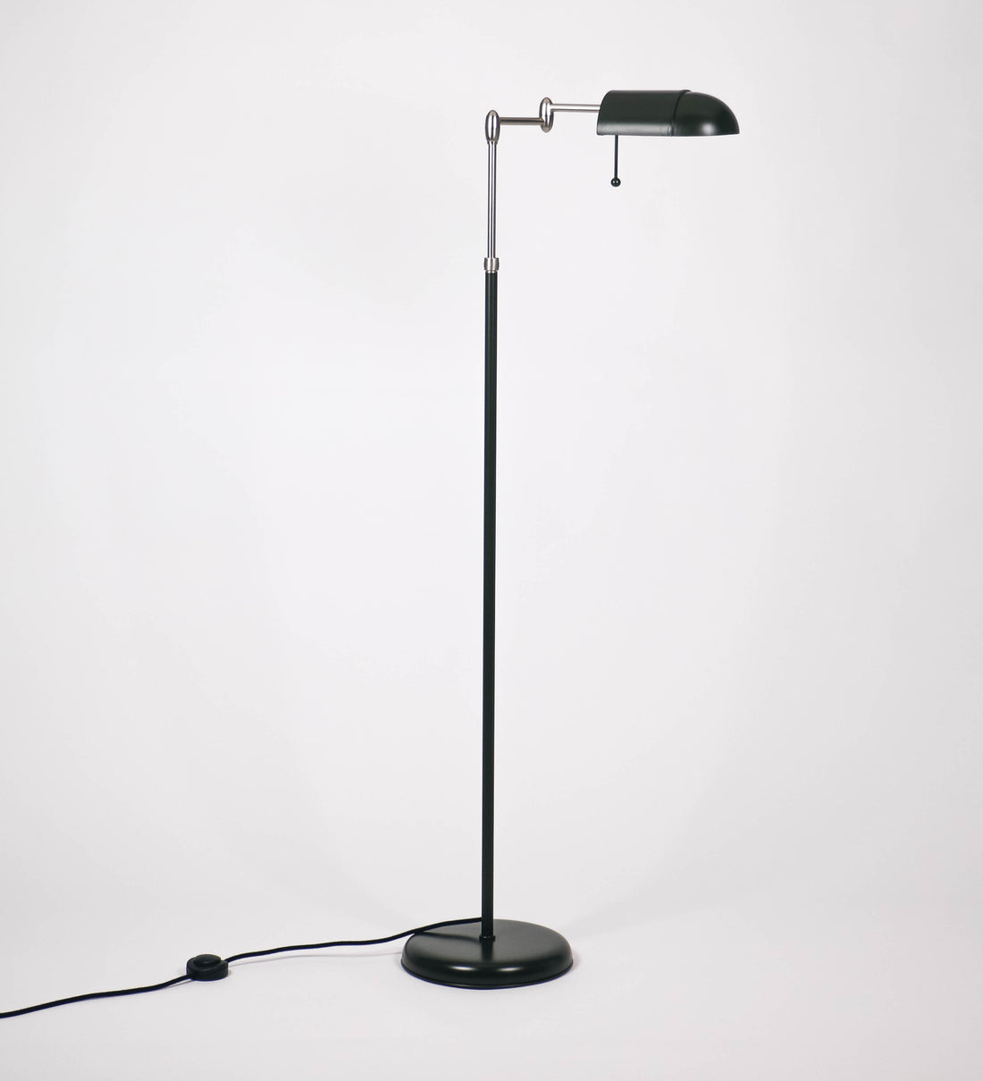 Candeeiro de pé com base circular em verde-azeitona. O corpo é fino e vertical, composto por uma haste verde-azeitona, e uma outra haste em níquel escovado, que permite ajustar a altura do candeeiro. A haste ajustável conecta a um braço horizontal, que sustenta o chapéu, e é manipulável. O chapéu é longo e arredondado, em verde-azeitona, e com uma pequena haste com uma esfera na ponta. O interior do chapéu é branco. O cabo é revestido têxtil em preto, com um interruptor de pé preto circular.