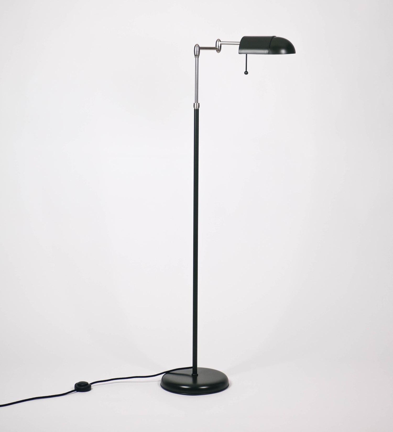 Candeeiro de pé com base circular em verde-azeitona. O corpo é fino e vertical, composto por uma haste verde-azeitona, e uma outra haste em níquel escovado, que permite ajustar a altura do candeeiro. A haste ajustável conecta a um braço horizontal, que sustenta o chapéu, e é manipulável. O chapéu é longo e arredondado, em verde-azeitona, e com uma pequena haste com uma esfera na ponta. O interior do chapéu é branco. O cabo é revestido têxtil em preto, com um interruptor de pé preto circular.