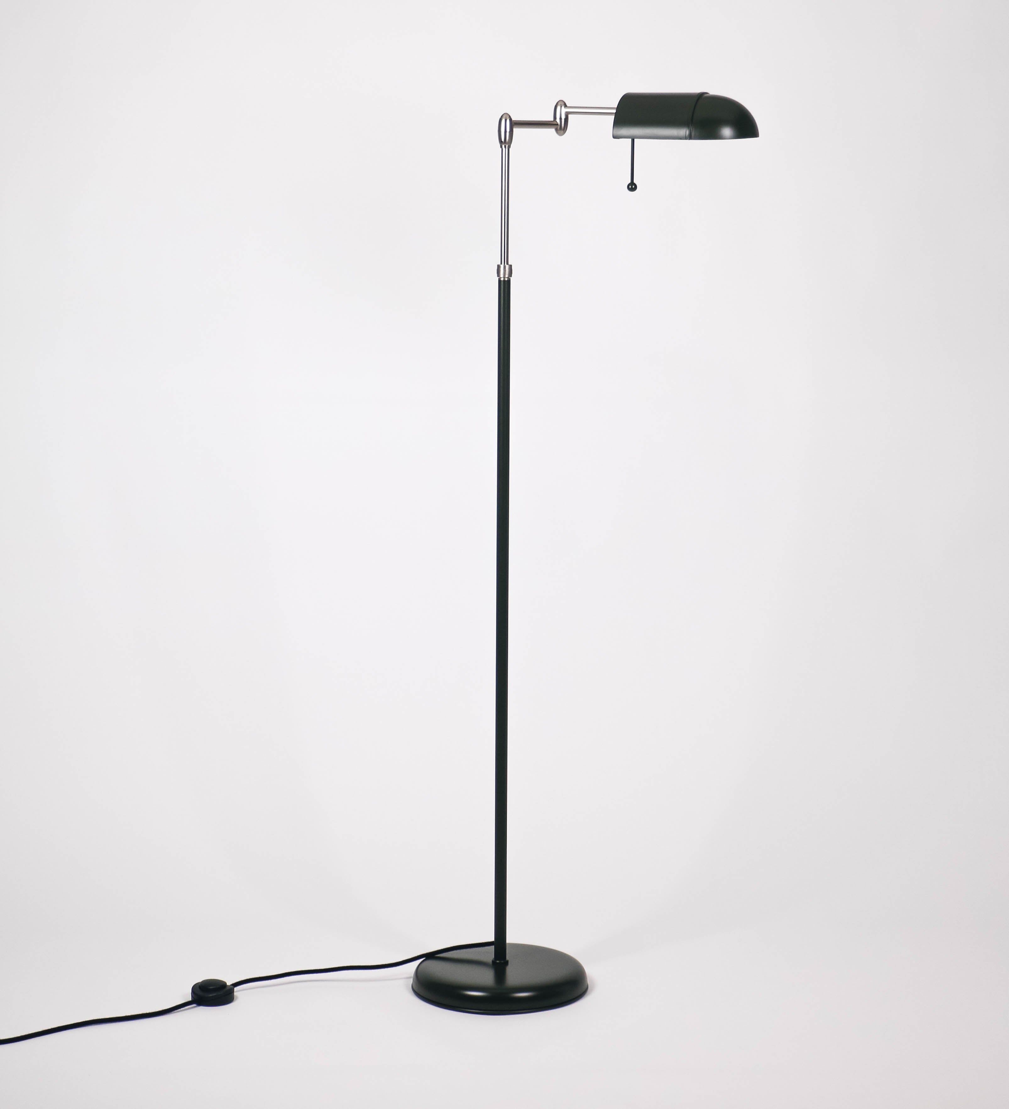 Candeeiro de pé com base circular em verde-azeitona. O corpo é fino e vertical, composto por uma haste verde-azeitona, e uma outra haste em níquel escovado, que permite ajustar a altura do candeeiro. A haste ajustável conecta a um braço horizontal, que sustenta o chapéu, e é manipulável. O chapéu é longo e arredondado, em verde-azeitona, e com uma pequena haste com uma esfera na ponta. O interior do chapéu é branco. O cabo é revestido têxtil em preto, com um interruptor de pé preto circular.