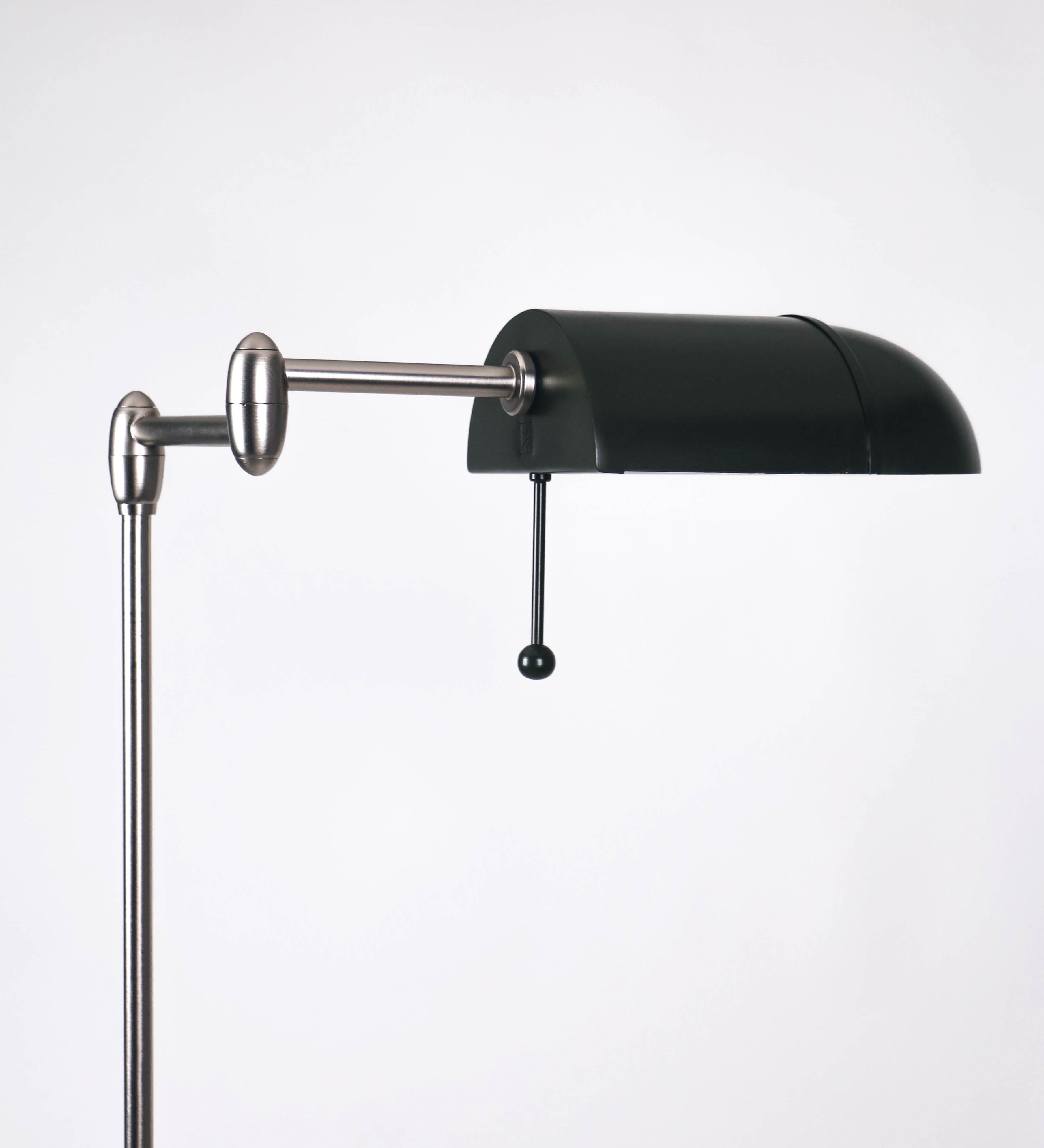 Candeeiro de pé com base circular em verde-azeitona. O corpo é fino e vertical, composto por uma haste verde-azeitona, e uma outra haste em níquel escovado, que permite ajustar a altura do candeeiro. A haste ajustável conecta a um braço horizontal, que sustenta o chapéu, e é manipulável. O chapéu é longo e arredondado, em verde-azeitona, e com uma pequena haste com uma esfera na ponta. O interior do chapéu é branco. O cabo é revestido têxtil em preto, com um interruptor de pé preto circular.