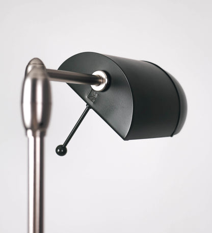 Candeeiro de pé com base circular em verde-azeitona. O corpo é fino e vertical, composto por uma haste verde-azeitona, e uma outra haste em níquel escovado, que permite ajustar a altura do candeeiro. A haste ajustável conecta a um braço horizontal, que sustenta o chapéu, e é manipulável. O chapéu é longo e arredondado, em verde-azeitona, e com uma pequena haste com uma esfera na ponta. O interior do chapéu é branco. O cabo é revestido têxtil em preto, com um interruptor de pé preto circular.