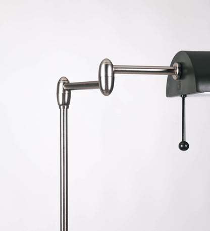 Candeeiro de pé com base circular em verde-azeitona. O corpo é fino e vertical, composto por uma haste verde-azeitona, e uma outra haste em níquel escovado, que permite ajustar a altura do candeeiro. A haste ajustável conecta a um braço horizontal, que sustenta o chapéu, e é manipulável. O chapéu é longo e arredondado, em verde-azeitona, e com uma pequena haste com uma esfera na ponta. O interior do chapéu é branco. O cabo é revestido têxtil em preto, com um interruptor de pé preto circular.