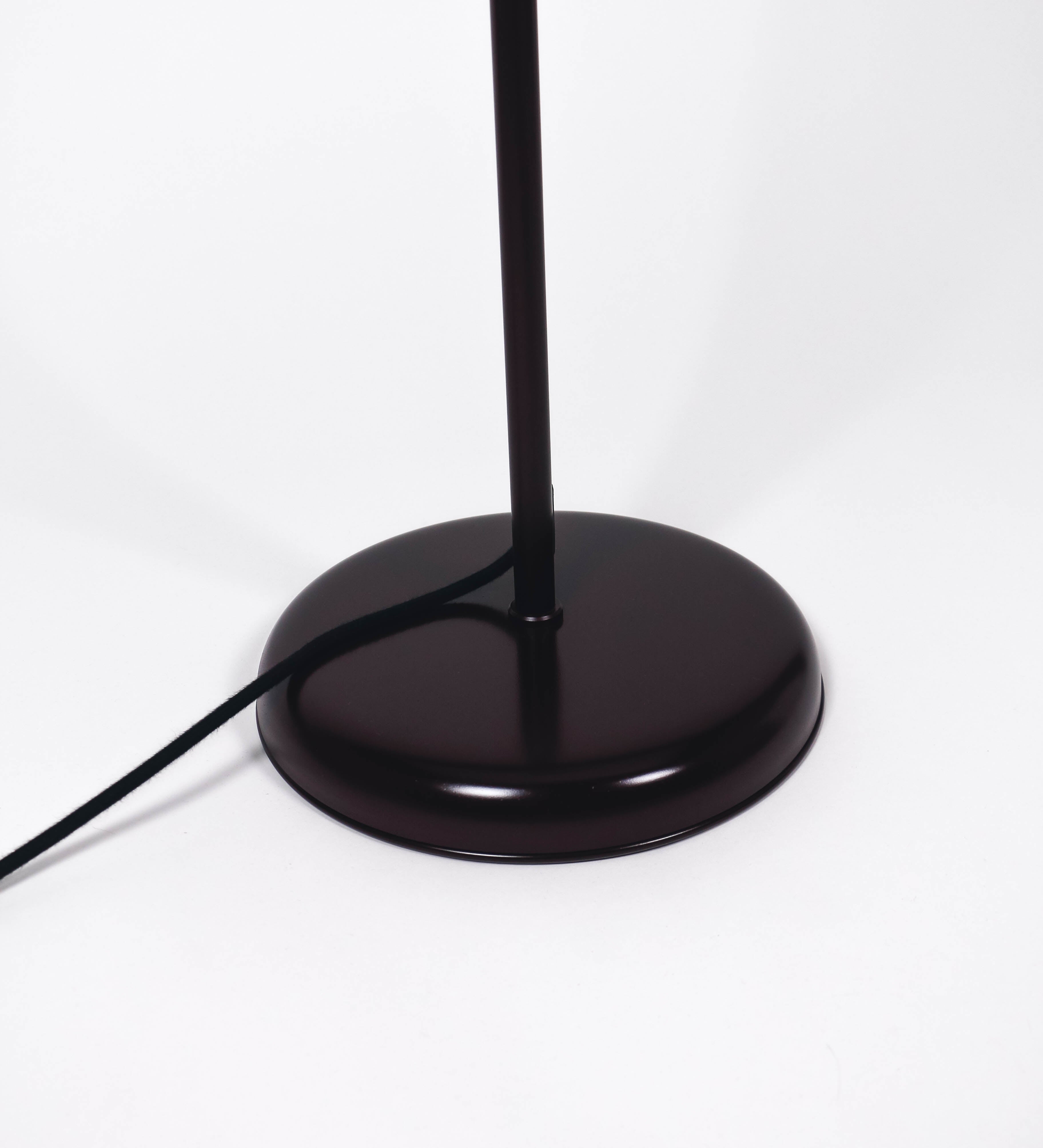 Candeeiro de pé com base circular em tom vinho. O corpo é fino e vertical, composto por uma haste tom vinho, e uma outra haste em latão escovado, que permite ajustar a altura do candeeiro. A haste ajustável conecta a um braço horizontal, que sustenta o chapéu, e é manipulável. O chapéu é longo e arredondado, em tom vinho, e com uma pequena haste com uma esfera na ponta. O interior do chapéu é branco. O cabo é revestido têxtil em preto, com um interruptor de pé preto circular.