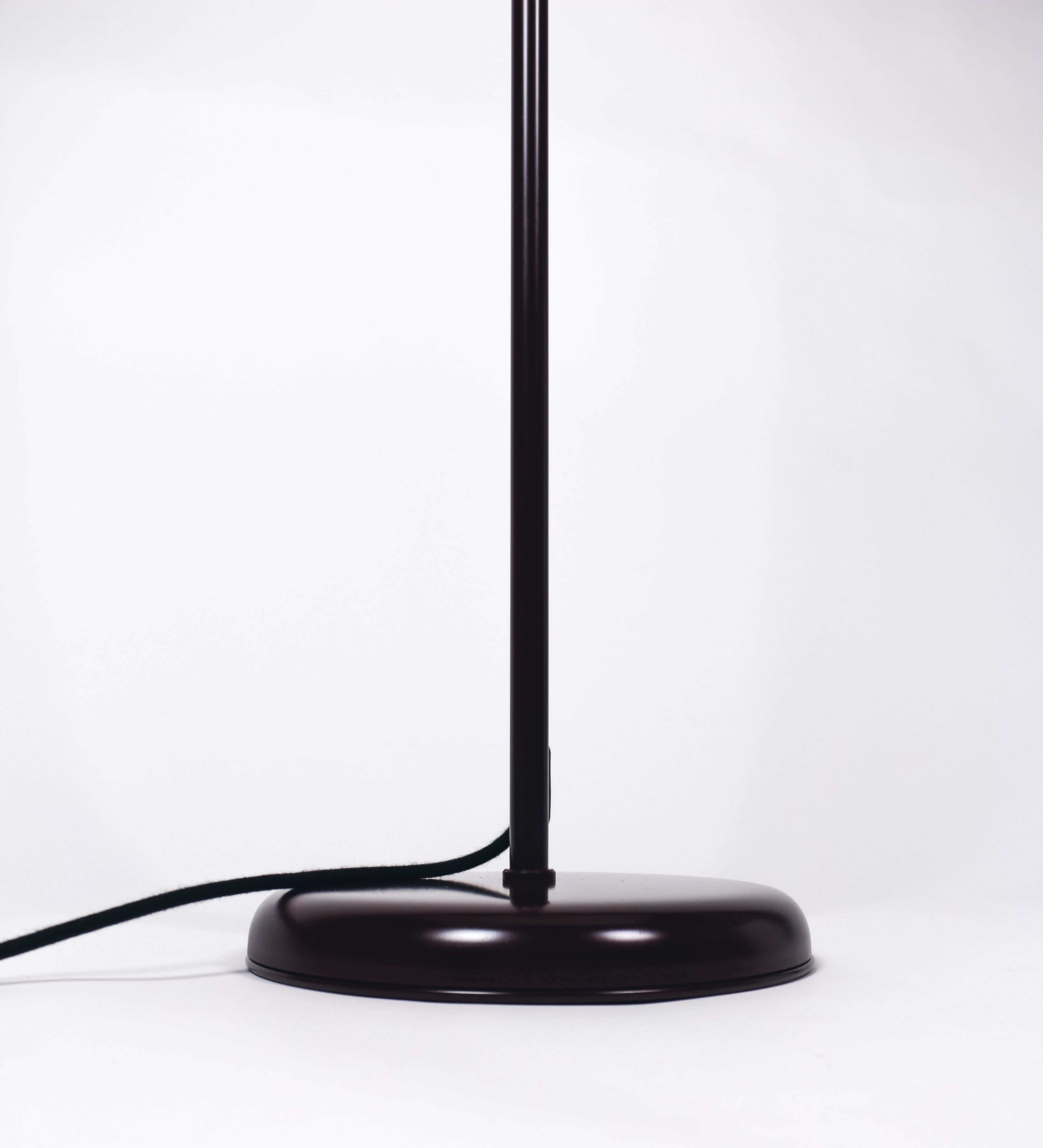 Candeeiro de pé com base circular em tom vinho. O corpo é fino e vertical, composto por uma haste tom vinho, e uma outra haste em latão escovado, que permite ajustar a altura do candeeiro. A haste ajustável conecta a um braço horizontal, que sustenta o chapéu, e é manipulável. O chapéu é longo e arredondado, em tom vinho, e com uma pequena haste com uma esfera na ponta. O interior do chapéu é branco. O cabo é revestido têxtil em preto, com um interruptor de pé preto circular.