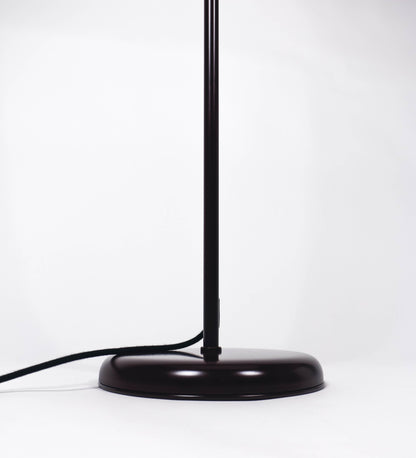 Candeeiro de pé com base circular em tom vinho. O corpo é fino e vertical, composto por uma haste tom vinho, e uma outra haste em latão escovado, que permite ajustar a altura do candeeiro. A haste ajustável conecta a um braço horizontal, que sustenta o chapéu, e é manipulável. O chapéu é longo e arredondado, em tom vinho, e com uma pequena haste com uma esfera na ponta. O interior do chapéu é branco. O cabo é revestido têxtil em preto, com um interruptor de pé preto circular.