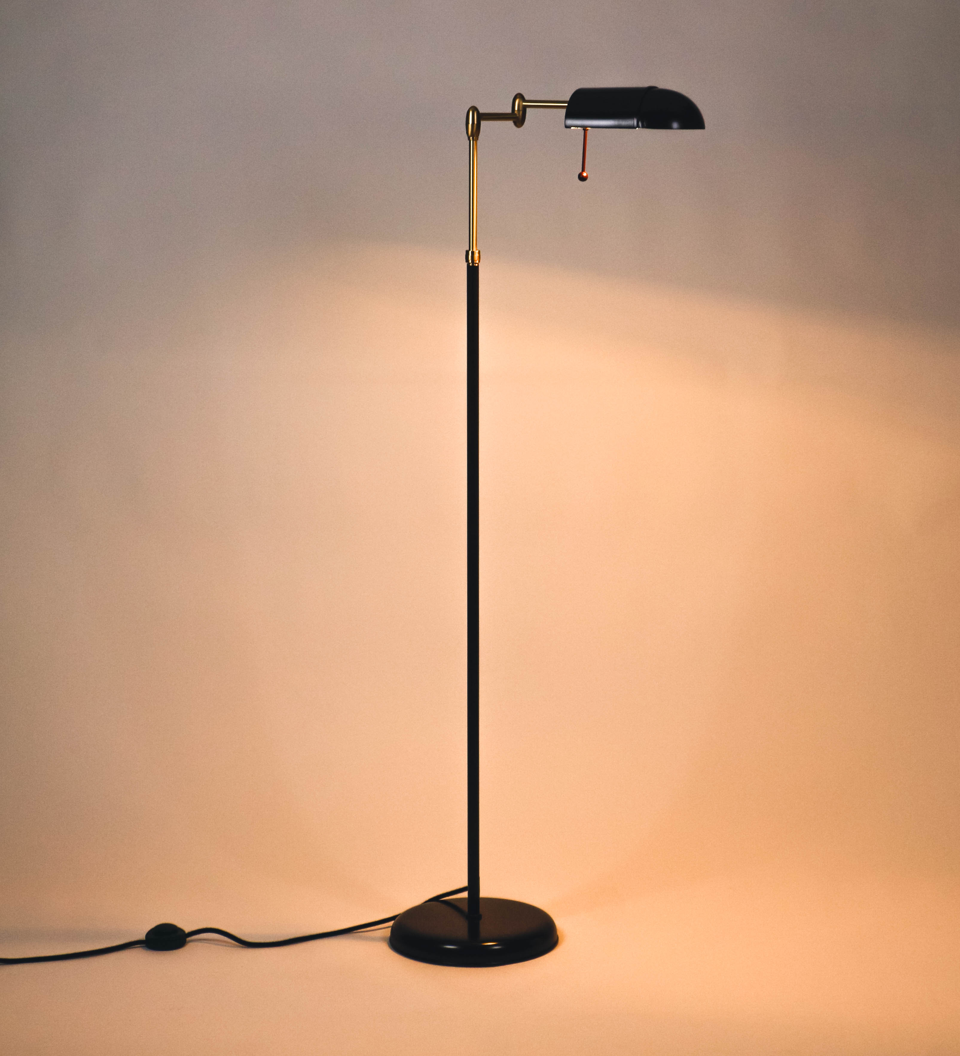 Candeeiro de pé com base circular em tom vinho. O corpo é fino e vertical, composto por uma haste tom vinho, e uma outra haste em latão escovado, que permite ajustar a altura do candeeiro. A haste ajustável conecta a um braço horizontal, que sustenta o chapéu, e é manipulável. O chapéu é longo e arredondado, em tom vinho, e com uma pequena haste com uma esfera na ponta. O interior do chapéu é branco. O cabo é revestido têxtil em preto, com um interruptor de pé preto circular.