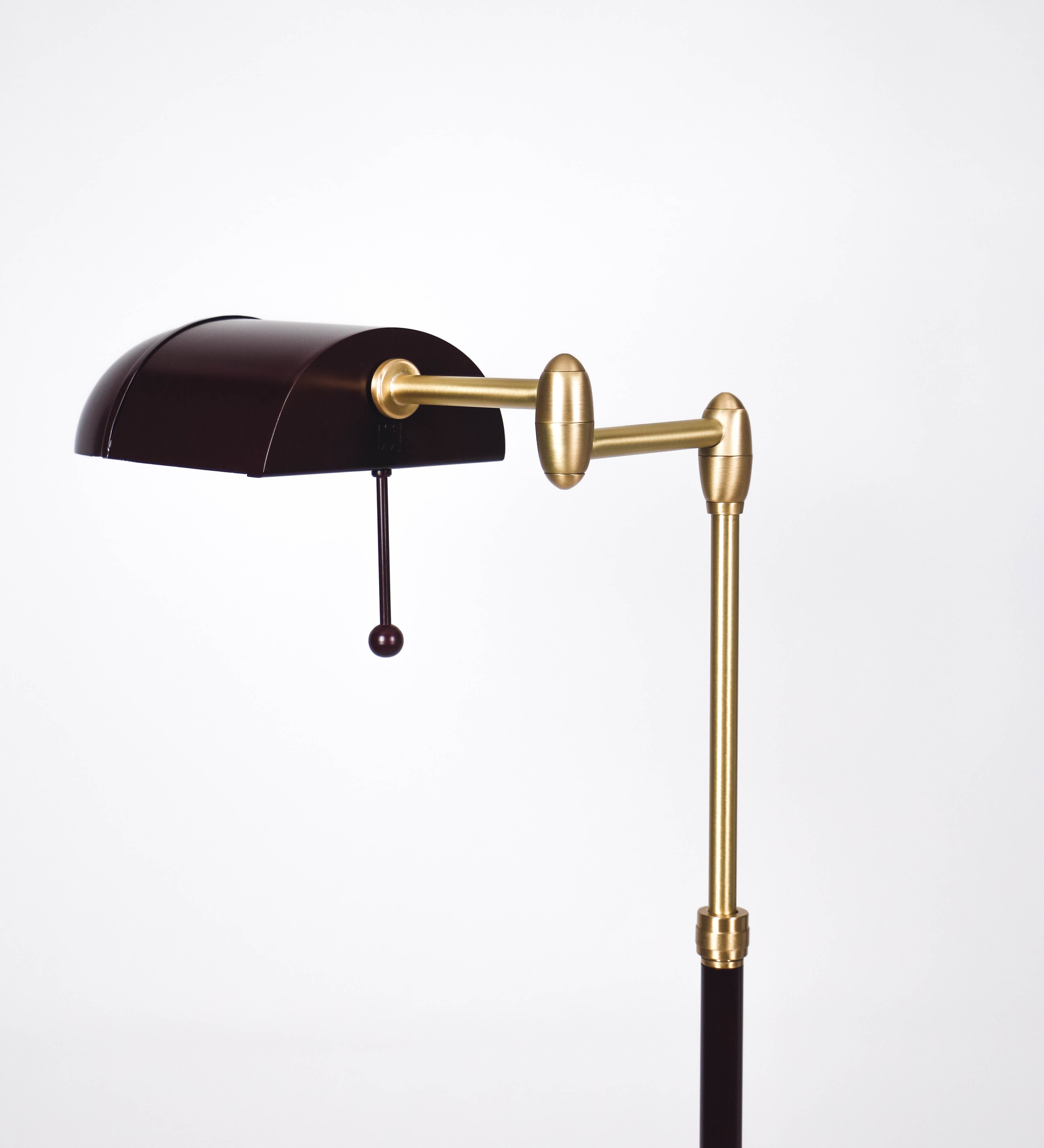 Candeeiro de pé com base circular em tom vinho. O corpo é fino e vertical, composto por uma haste tom vinho, e uma outra haste em latão escovado, que permite ajustar a altura do candeeiro. A haste ajustável conecta a um braço horizontal, que sustenta o chapéu, e é manipulável. O chapéu é longo e arredondado, em tom vinho, e com uma pequena haste com uma esfera na ponta. O interior do chapéu é branco. O cabo é revestido têxtil em preto, com um interruptor de pé preto circular.