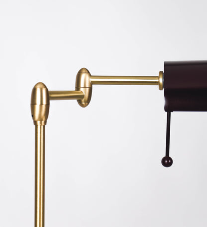 Candeeiro de pé com base circular em tom vinho. O corpo é fino e vertical, composto por uma haste tom vinho, e uma outra haste em latão escovado, que permite ajustar a altura do candeeiro. A haste ajustável conecta a um braço horizontal, que sustenta o chapéu, e é manipulável. O chapéu é longo e arredondado, em tom vinho, e com uma pequena haste com uma esfera na ponta. O interior do chapéu é branco. O cabo é revestido têxtil em preto, com um interruptor de pé preto circular.