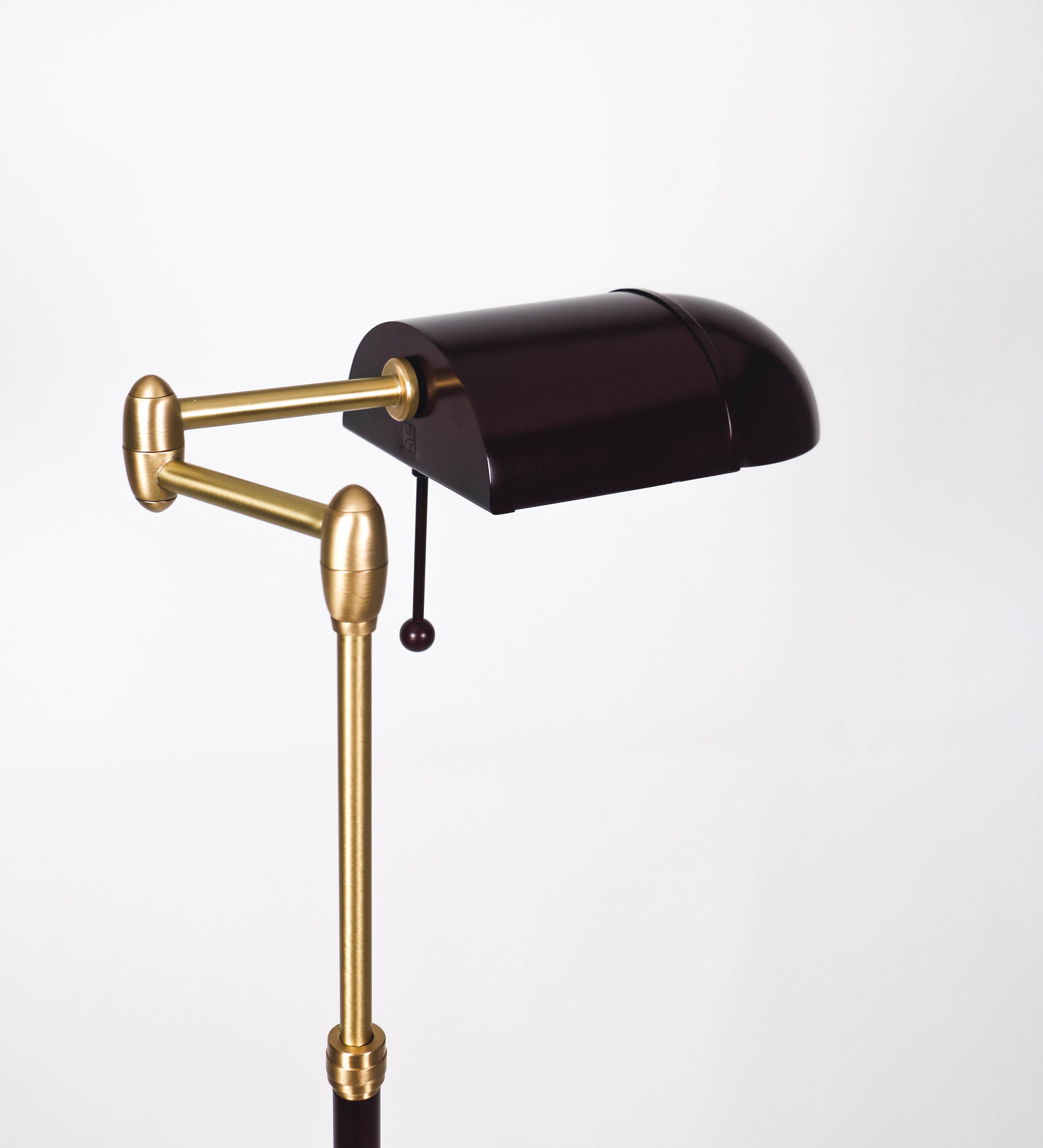 Candeeiro de pé com base circular em tom vinho. O corpo é fino e vertical, composto por uma haste tom vinho, e uma outra haste em latão escovado, que permite ajustar a altura do candeeiro. A haste ajustável conecta a um braço horizontal, que sustenta o chapéu, e é manipulável. O chapéu é longo e arredondado, em tom vinho, e com uma pequena haste com uma esfera na ponta. O interior do chapéu é branco. O cabo é revestido têxtil em preto, com um interruptor de pé preto circular.