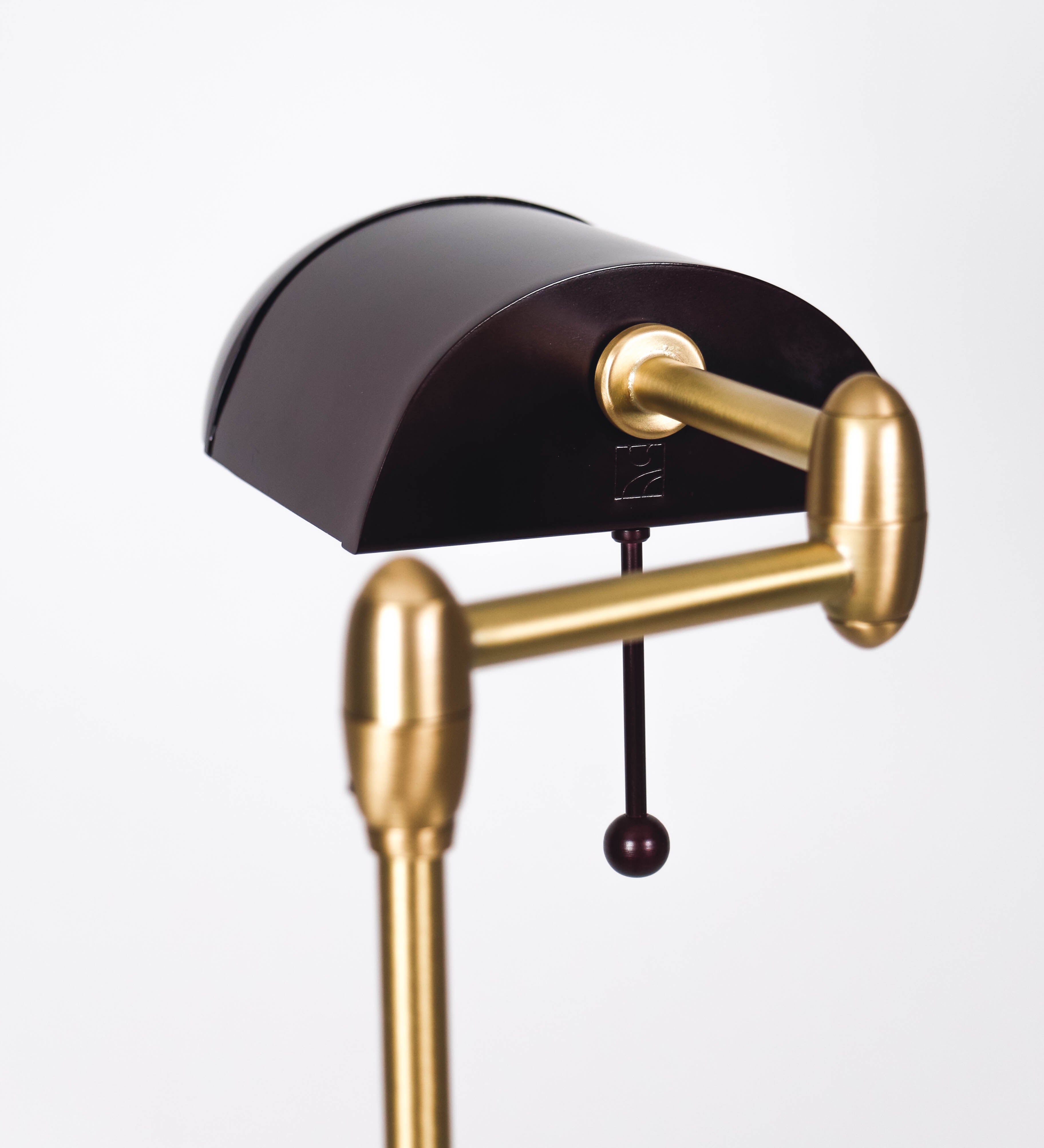 Candeeiro de pé com base circular em tom vinho. O corpo é fino e vertical, composto por uma haste tom vinho, e uma outra haste em latão escovado, que permite ajustar a altura do candeeiro. A haste ajustável conecta a um braço horizontal, que sustenta o chapéu, e é manipulável. O chapéu é longo e arredondado, em tom vinho, e com uma pequena haste com uma esfera na ponta. O interior do chapéu é branco. O cabo é revestido têxtil em preto, com um interruptor de pé preto circular.
