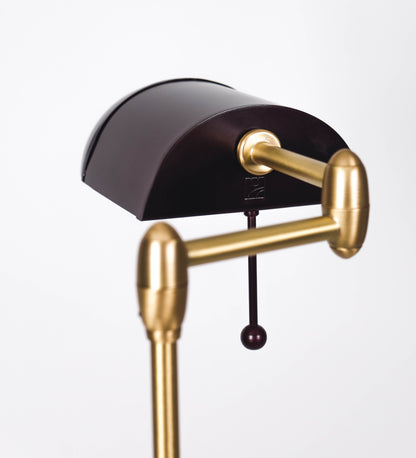 Candeeiro de pé com base circular em tom vinho. O corpo é fino e vertical, composto por uma haste tom vinho, e uma outra haste em latão escovado, que permite ajustar a altura do candeeiro. A haste ajustável conecta a um braço horizontal, que sustenta o chapéu, e é manipulável. O chapéu é longo e arredondado, em tom vinho, e com uma pequena haste com uma esfera na ponta. O interior do chapéu é branco. O cabo é revestido têxtil em preto, com um interruptor de pé preto circular.