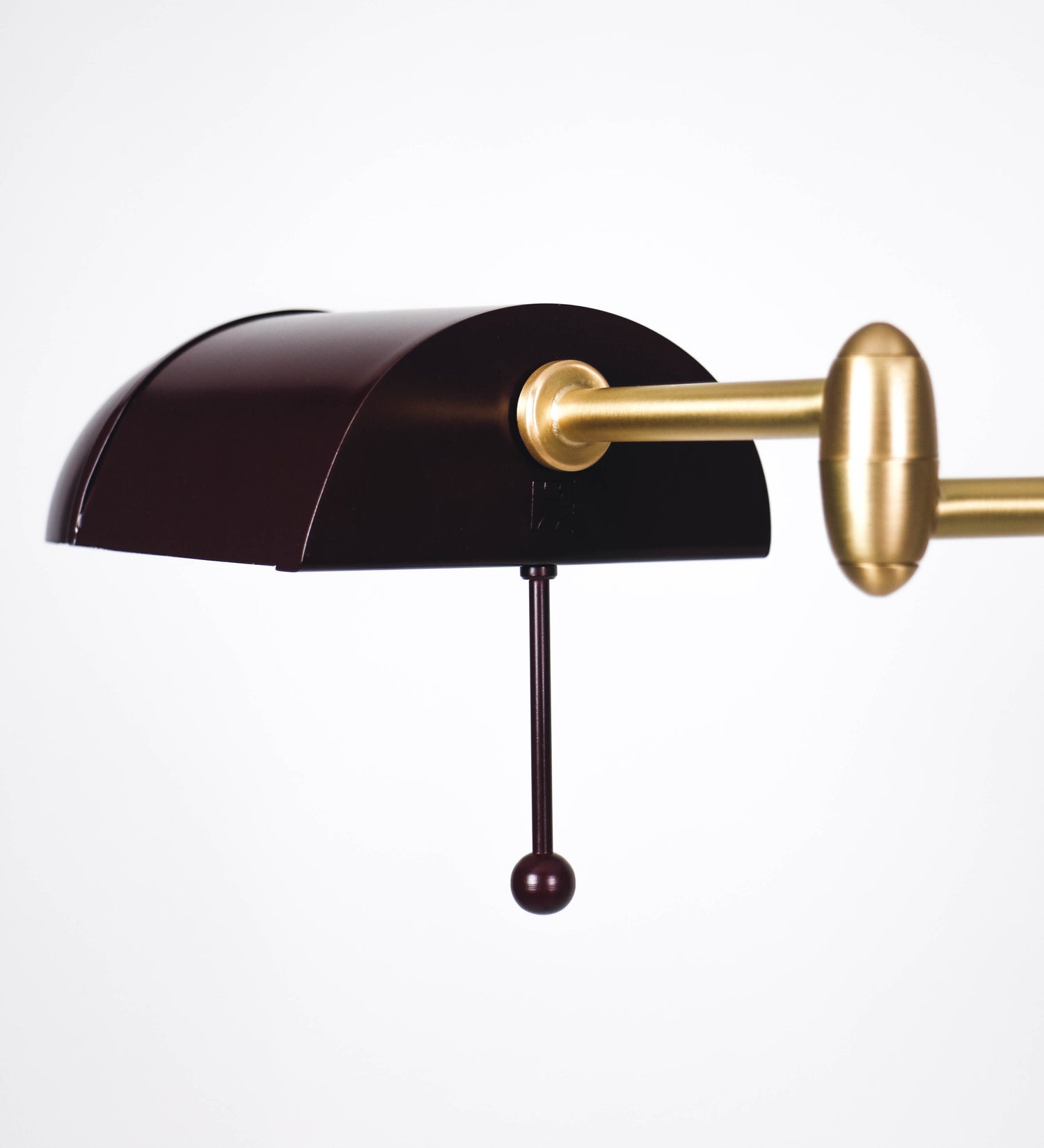 Candeeiro de pé com base circular em tom vinho. O corpo é fino e vertical, composto por uma haste tom vinho, e uma outra haste em latão escovado, que permite ajustar a altura do candeeiro. A haste ajustável conecta a um braço horizontal, que sustenta o chapéu, e é manipulável. O chapéu é longo e arredondado, em tom vinho, e com uma pequena haste com uma esfera na ponta. O interior do chapéu é branco. O cabo é revestido têxtil em preto, com um interruptor de pé preto circular.