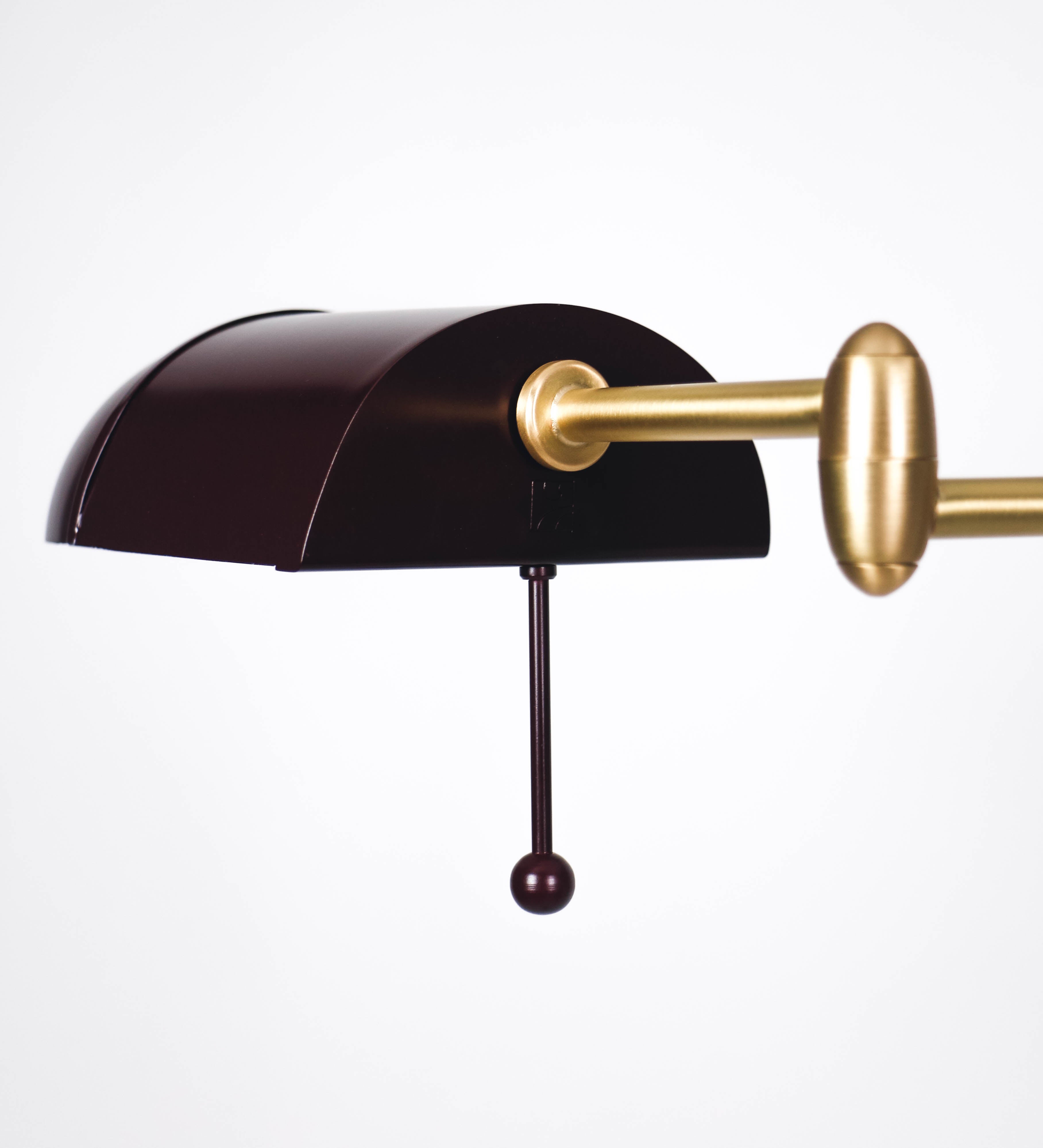 Candeeiro de pé com base circular em tom vinho. O corpo é fino e vertical, composto por uma haste tom vinho, e uma outra haste em latão escovado, que permite ajustar a altura do candeeiro. A haste ajustável conecta a um braço horizontal, que sustenta o chapéu, e é manipulável. O chapéu é longo e arredondado, em tom vinho, e com uma pequena haste com uma esfera na ponta. O interior do chapéu é branco. O cabo é revestido têxtil em preto, com um interruptor de pé preto circular.