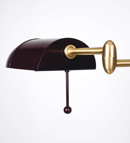 Candeeiro de pé com base circular em tom vinho. O corpo é fino e vertical, composto por uma haste tom vinho, e uma outra haste em latão escovado, que permite ajustar a altura do candeeiro. A haste ajustável conecta a um braço horizontal, que sustenta o chapéu, e é manipulável. O chapéu é longo e arredondado, em tom vinho, e com uma pequena haste com uma esfera na ponta. O interior do chapéu é branco. O cabo é revestido têxtil em preto, com um interruptor de pé preto circular.