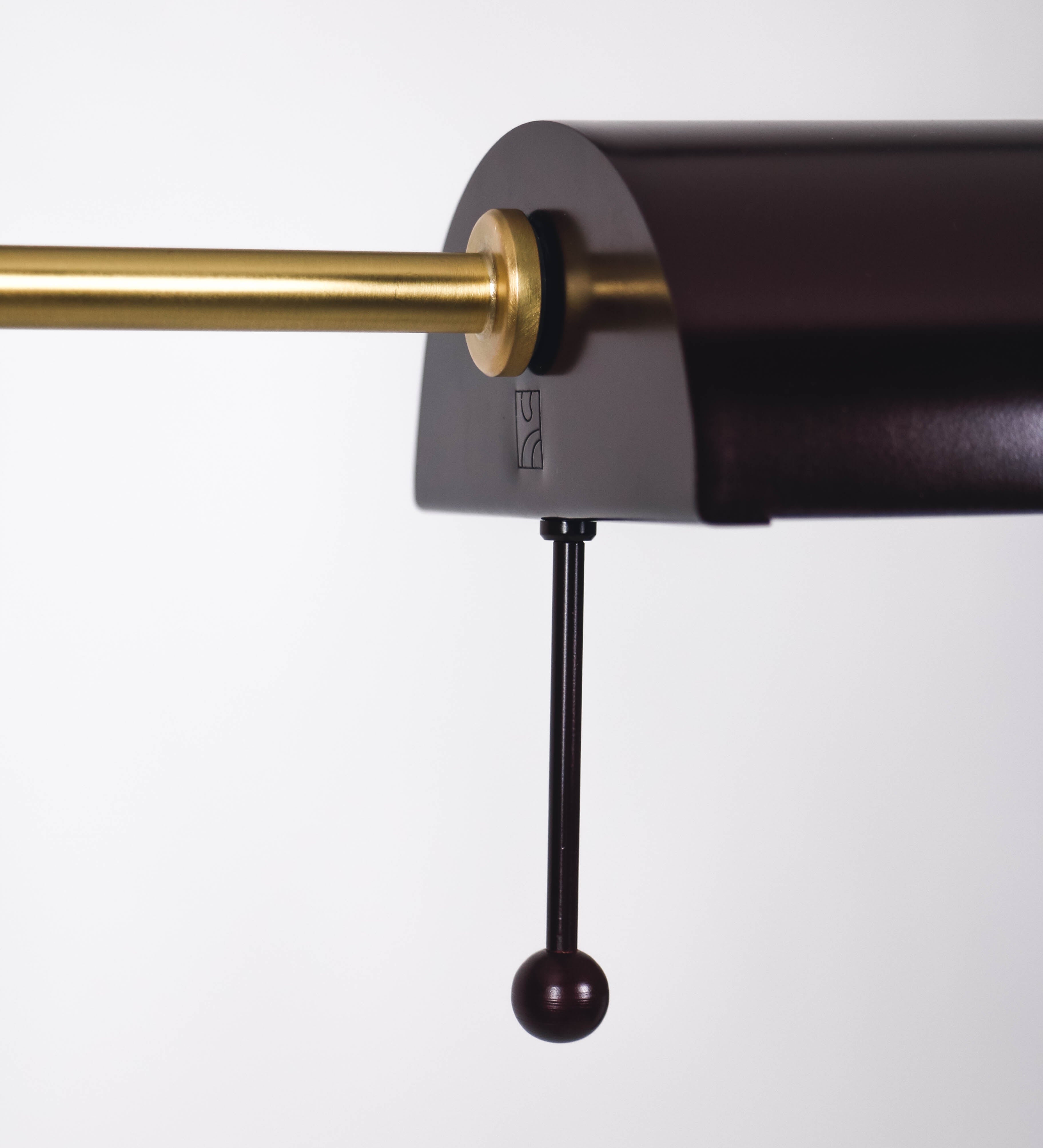 Candeeiro de pé com base circular em tom vinho. O corpo é fino e vertical, composto por uma haste tom vinho, e uma outra haste em latão escovado, que permite ajustar a altura do candeeiro. A haste ajustável conecta a um braço horizontal, que sustenta o chapéu, e é manipulável. O chapéu é longo e arredondado, em tom vinho, e com uma pequena haste com uma esfera na ponta. O interior do chapéu é branco. O cabo é revestido têxtil em preto, com um interruptor de pé preto circular.