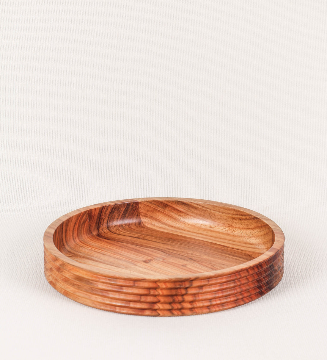 Fruteira com um design minimalista e distinto. A fruteira é de madeira de Acácia-Austrália, com uma tonalidade escura e verniz foodsafe semi-brilhante. A peça é de formato circular e com uma borda de cerca de 3 cm de altura, decorada com relevos lineares dispostos horizontalmente, tipo ranhuras, que envolvem a fruteira.