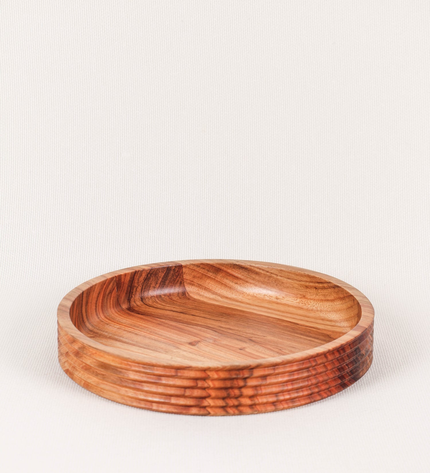 Fruteira com um design minimalista e distinto. A fruteira é de madeira de Acácia-Austrália, com uma tonalidade escura e verniz foodsafe semi-brilhante. A peça é de formato circular e com uma borda de cerca de 3 cm de altura, decorada com relevos lineares dispostos horizontalmente, tipo ranhuras, que envolvem a fruteira.