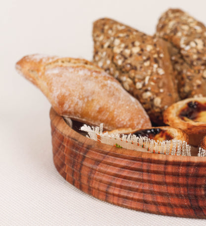 Fruteira com um design minimalista e distinto. A fruteira é de madeira de Acácia-Austrália, com uma tonalidade escura e verniz foodsafe semi-brilhante. A peça é de formato circular e com uma borda de cerca de 3 cm de altura, decorada com relevos lineares dispostos horizontalmente, tipo ranhuras, que envolvem a fruteira.