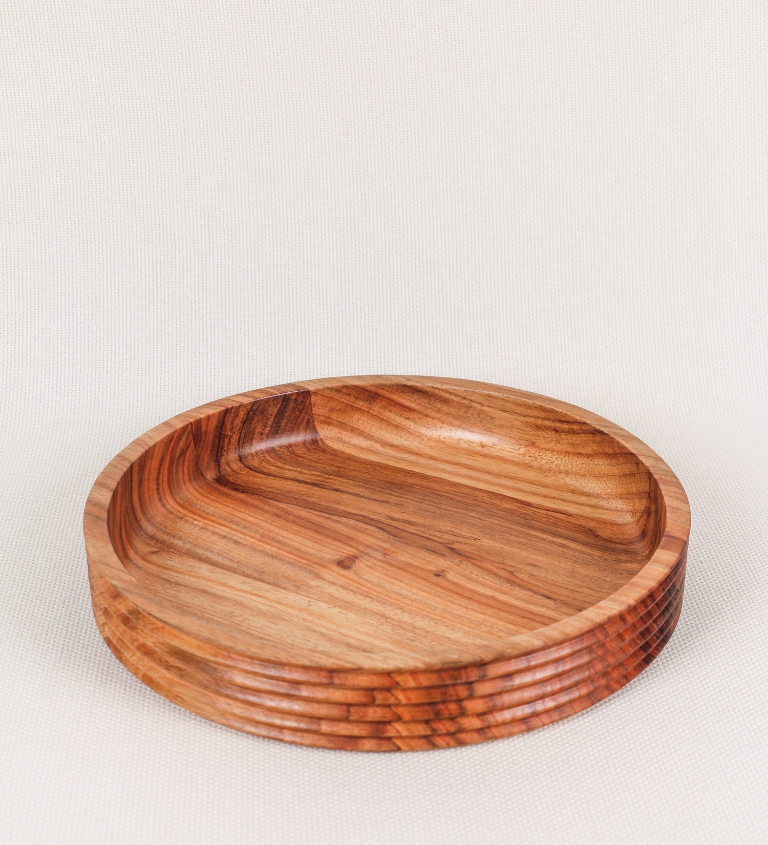 Fruteira com um design minimalista e distinto. A fruteira é de madeira de Acácia-Austrália, com uma tonalidade escura e verniz foodsafe semi-brilhante. A peça é de formato circular e com uma borda de cerca de 3 cm de altura, decorada com relevos lineares dispostos horizontalmente, tipo ranhuras, que envolvem a fruteira.