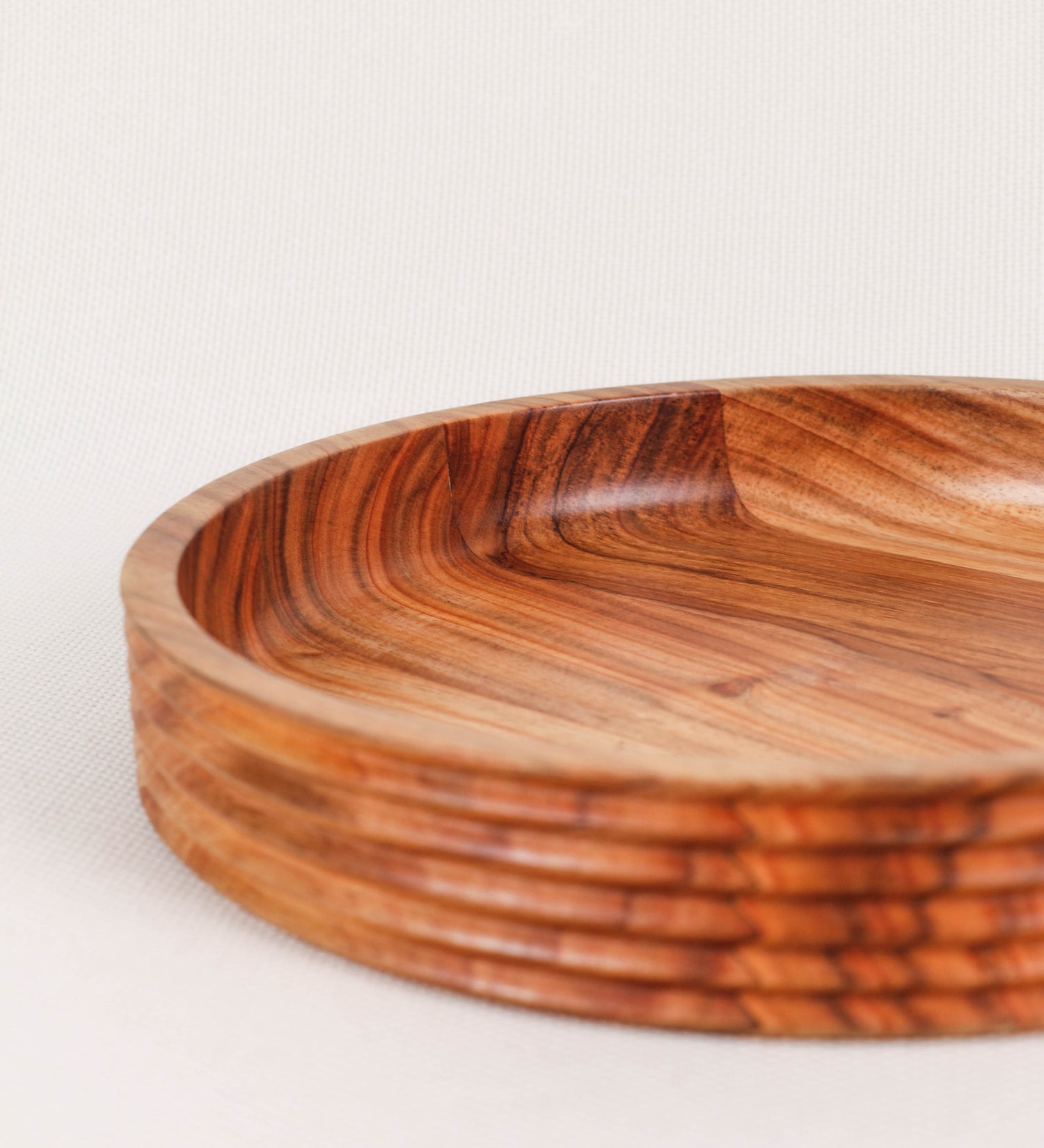 Fruteira com um design minimalista e distinto. A fruteira é de madeira de Acácia-Austrália, com uma tonalidade escura e verniz foodsafe semi-brilhante. A peça é de formato circular e com uma borda de cerca de 3 cm de altura, decorada com relevos lineares dispostos horizontalmente, tipo ranhuras, que envolvem a fruteira.