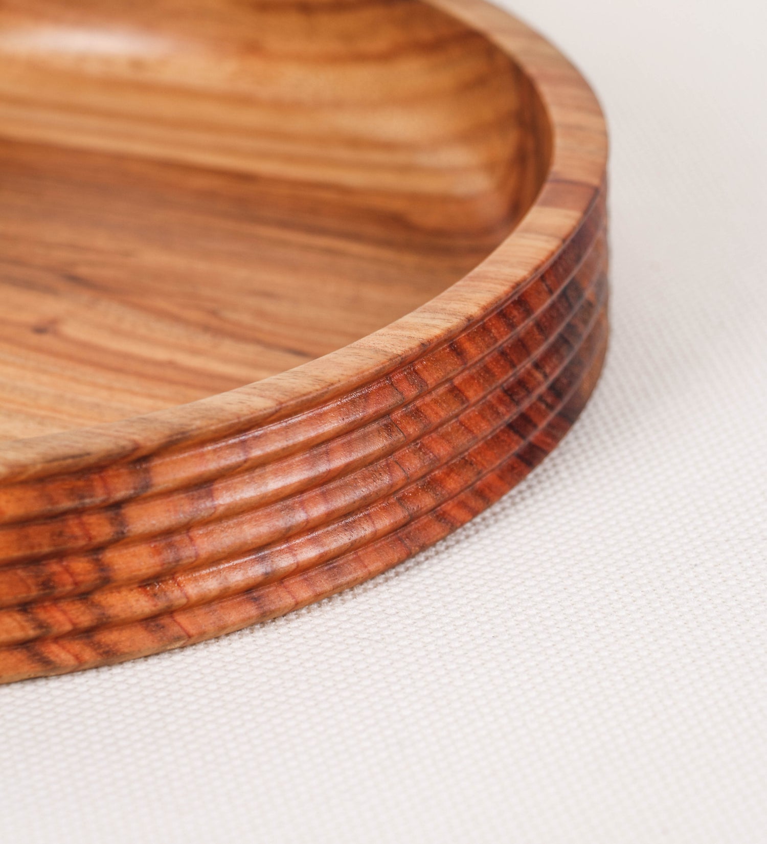 Fruteira com um design minimalista e distinto. A fruteira é de madeira de Acácia-Austrália, com uma tonalidade escura e verniz foodsafe semi-brilhante. A peça é de formato circular e com uma borda de cerca de 3 cm de altura, decorada com relevos lineares dispostos horizontalmente, tipo ranhuras, que envolvem a fruteira.