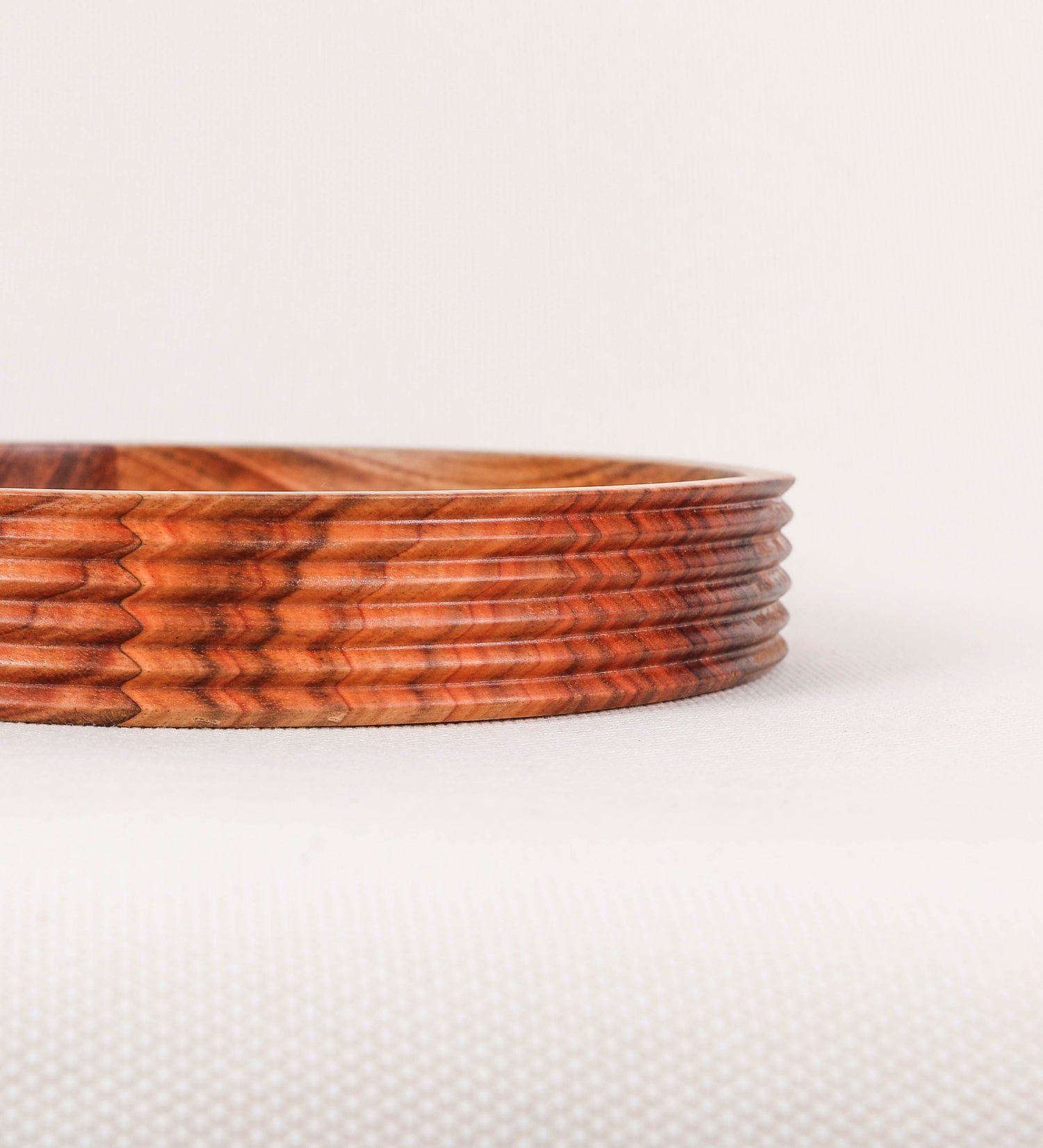 Fruteira com um design minimalista e distinto. A fruteira é de madeira de Acácia-Austrália, com uma tonalidade escura e verniz foodsafe semi-brilhante. A peça é de formato circular e com uma borda de cerca de 3 cm de altura, decorada com relevos lineares dispostos horizontalmente, tipo ranhuras, que envolvem a fruteira.