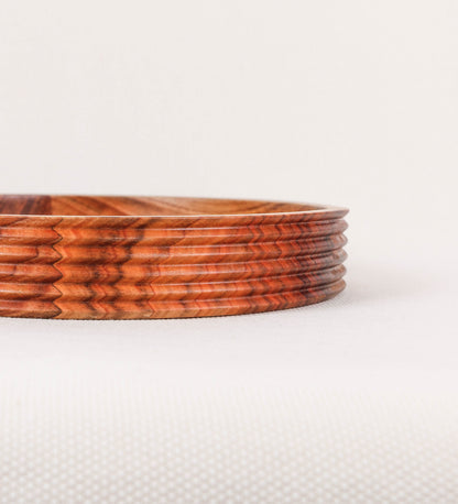 Fruteira com um design minimalista e distinto. A fruteira é de madeira de Acácia-Austrália, com uma tonalidade escura e verniz foodsafe semi-brilhante. A peça é de formato circular e com uma borda de cerca de 3 cm de altura, decorada com relevos lineares dispostos horizontalmente, tipo ranhuras, que envolvem a fruteira.