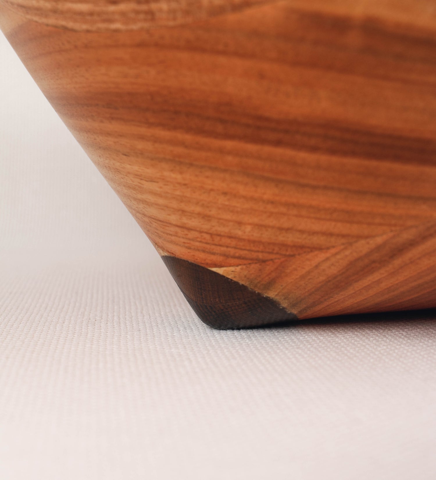 Fruteira com um design minimalista e distinto. A fruteira é de madeira de Acácia-Austrália, com uma tonalidade escura e verniz foodsafe semi-brilhante. A peça é de formato cónico e com um toque liso, semelhante a um pião, com uma ponteira em madeira também de tom mais escuro, quase negro. Apresenta ainda uma borda de cerca de 3 cm de altura, próxima da abertura da peça, decorada com relevos lineares dispostos horizontalmente, tipo ranhuras, que envolvem a fruteira.