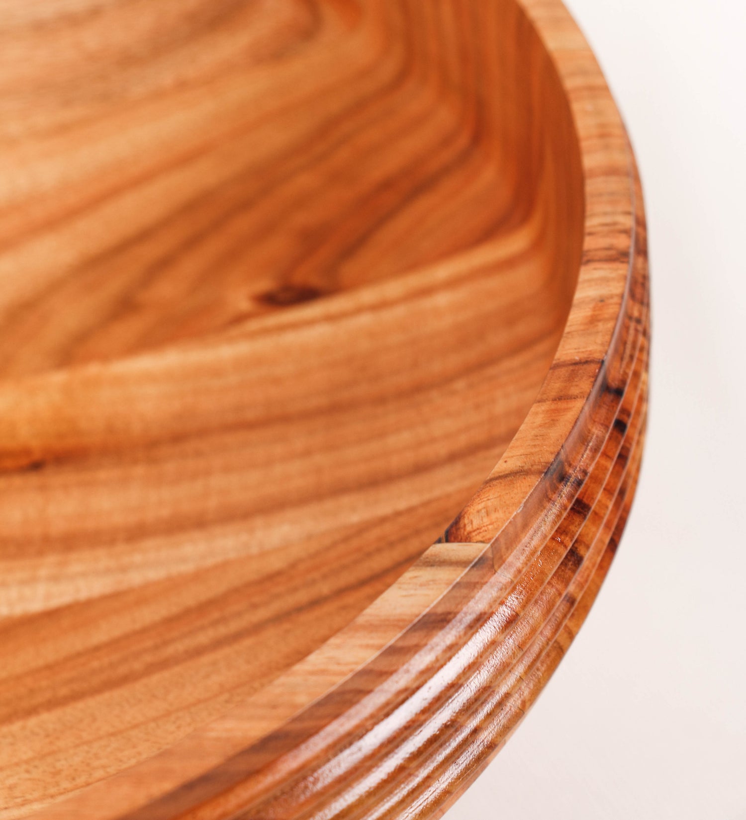 Fruteira com um design minimalista e distinto. A fruteira é de madeira de Acácia-Austrália, com uma tonalidade escura e verniz foodsafe semi-brilhante. A peça é de formato cónico e com um toque liso, semelhante a um pião, com uma ponteira em madeira também de tom mais escuro, quase negro. Apresenta ainda uma borda de cerca de 3 cm de altura, próxima da abertura da peça, decorada com relevos lineares dispostos horizontalmente, tipo ranhuras, que envolvem a fruteira.