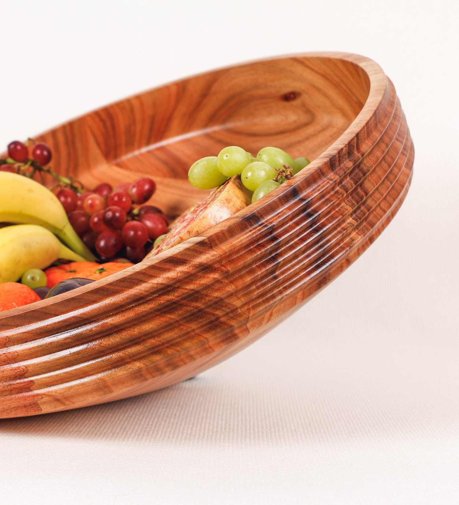 Fruteira com um design minimalista e distinto. A fruteira é de madeira de Acácia-Austrália, com uma tonalidade escura e verniz foodsafe semi-brilhante. A peça é de formato cónico e com um toque liso, semelhante a um pião, com uma ponteira em madeira também de tom mais escuro, quase negro. Apresenta ainda uma borda de cerca de 3 cm de altura, próxima da abertura da peça, decorada com relevos lineares dispostos horizontalmente, tipo ranhuras, que envolvem a fruteira.