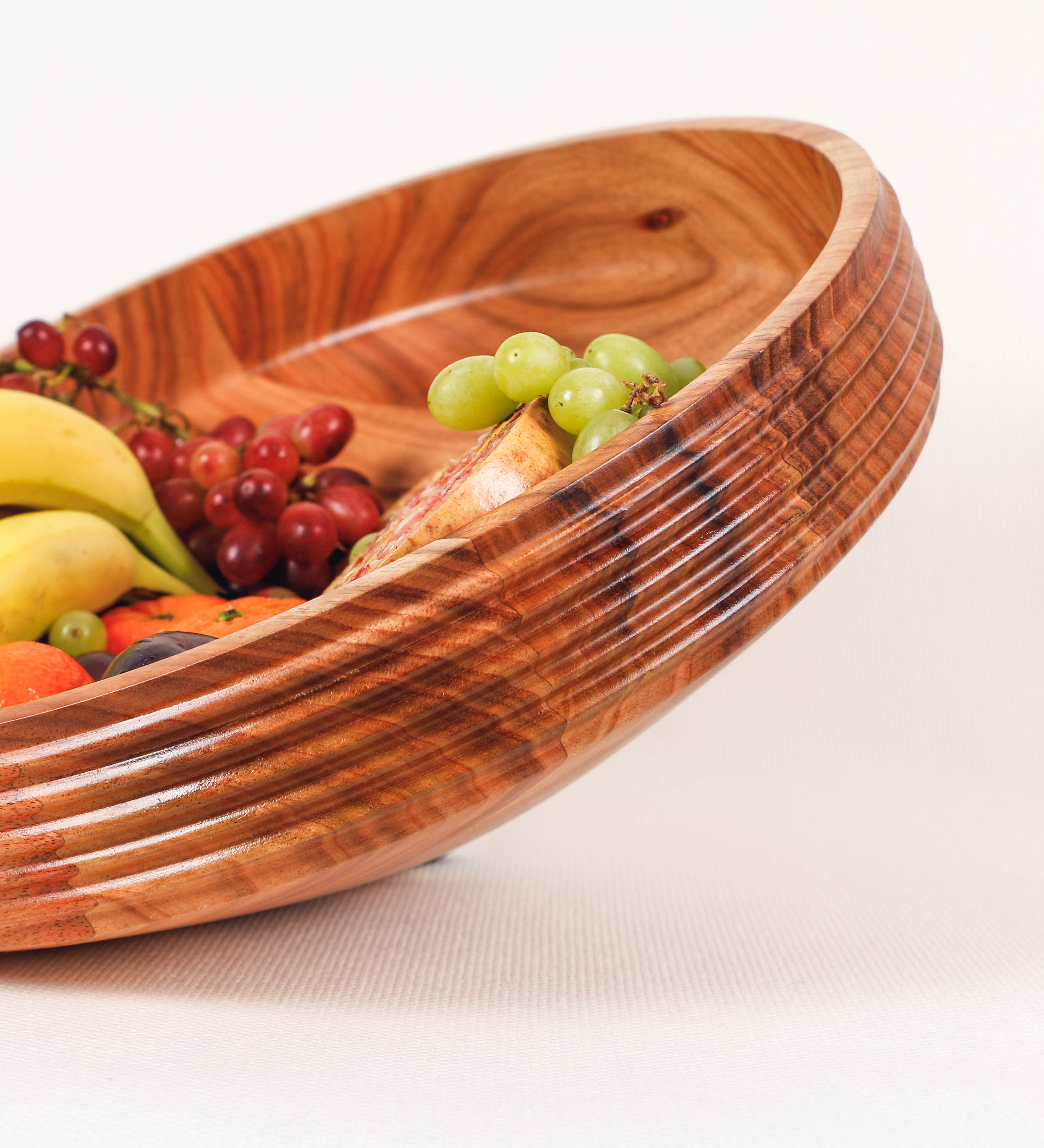 Fruteira com um design minimalista e distinto. A fruteira é de madeira de Acácia-Austrália, com uma tonalidade escura e verniz foodsafe semi-brilhante. A peça é de formato cónico e com um toque liso, semelhante a um pião, com uma ponteira em madeira também de tom mais escuro, quase negro. Apresenta ainda uma borda de cerca de 3 cm de altura, próxima da abertura da peça, decorada com relevos lineares dispostos horizontalmente, tipo ranhuras, que envolvem a fruteira.