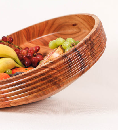 Fruteira com um design minimalista e distinto. A fruteira é de madeira de Acácia-Austrália, com uma tonalidade escura e verniz foodsafe semi-brilhante. A peça é de formato cónico e com um toque liso, semelhante a um pião, com uma ponteira em madeira também de tom mais escuro, quase negro. Apresenta ainda uma borda de cerca de 3 cm de altura, próxima da abertura da peça, decorada com relevos lineares dispostos horizontalmente, tipo ranhuras, que envolvem a fruteira.