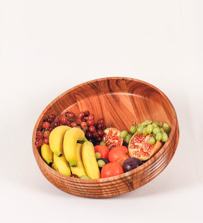 Fruteira com um design minimalista e distinto. A fruteira é de madeira de Acácia-Austrália, com uma tonalidade escura e verniz foodsafe semi-brilhante. A peça é de formato cónico e com um toque liso, semelhante a um pião, com uma ponteira em madeira também de tom mais escuro, quase negro. Apresenta ainda uma borda de cerca de 3 cm de altura, próxima da abertura da peça, decorada com relevos lineares dispostos horizontalmente, tipo ranhuras, que envolvem a fruteira.