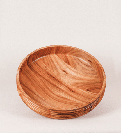 Fruteira com um design minimalista e distinto. A fruteira é de madeira de Acácia-Austrália, com uma tonalidade escura e verniz foodsafe semi-brilhante. A peça é de formato cónico e com um toque liso, semelhante a um pião, com uma ponteira em madeira também de tom mais escuro, quase negro. Apresenta ainda uma borda de cerca de 3 cm de altura, próxima da abertura da peça, decorada com relevos lineares dispostos horizontalmente, tipo ranhuras, que envolvem a fruteira.