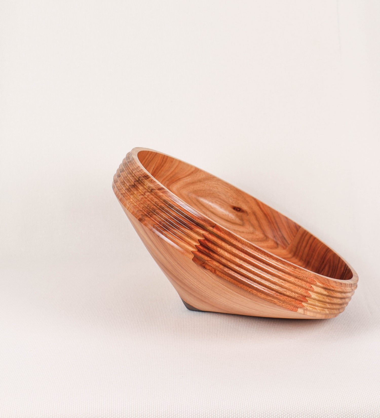 Fruteira com um design minimalista e distinto. A fruteira é de madeira de Acácia-Austrália, com uma tonalidade escura e verniz foodsafe semi-brilhante. A peça é de formato cónico e com um toque liso, semelhante a um pião, com uma ponteira em madeira também de tom mais escuro, quase negro. Apresenta ainda uma borda de cerca de 3 cm de altura, próxima da abertura da peça, decorada com relevos lineares dispostos horizontalmente, tipo ranhuras, que envolvem a fruteira.