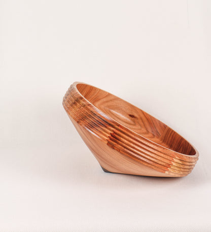 Fruteira com um design minimalista e distinto. A fruteira é de madeira de Acácia-Austrália, com uma tonalidade escura e verniz foodsafe semi-brilhante. A peça é de formato cónico e com um toque liso, semelhante a um pião, com uma ponteira em madeira também de tom mais escuro, quase negro. Apresenta ainda uma borda de cerca de 3 cm de altura, próxima da abertura da peça, decorada com relevos lineares dispostos horizontalmente, tipo ranhuras, que envolvem a fruteira.