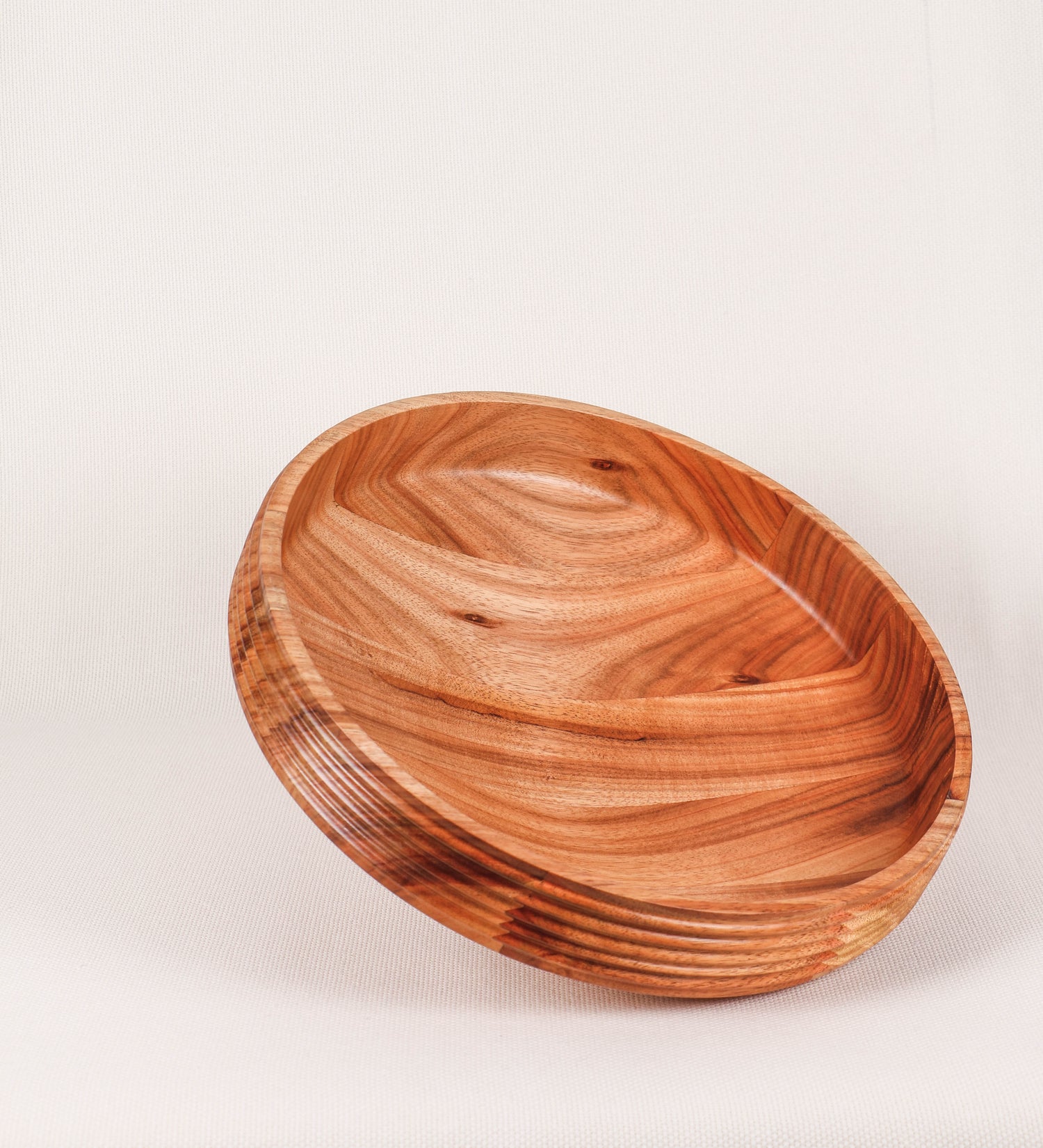 Fruteira com um design minimalista e distinto. A fruteira é de madeira de Acácia-Austrália, com uma tonalidade escura e verniz foodsafe semi-brilhante. A peça é de formato cónico e com um toque liso, semelhante a um pião, com uma ponteira em madeira também de tom mais escuro, quase negro. Apresenta ainda uma borda de cerca de 3 cm de altura, próxima da abertura da peça, decorada com relevos lineares dispostos horizontalmente, tipo ranhuras, que envolvem a fruteira.