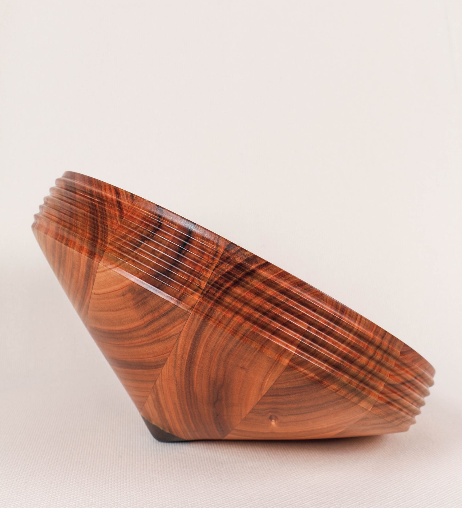 Fruteira com um design minimalista e distinto. A fruteira é de madeira de Acácia-Austrália, com uma tonalidade escura e verniz foodsafe semi-brilhante. A peça é de formato cónico e com um toque liso, semelhante a um pião, com uma ponteira em madeira também de tom mais escuro, quase negro. Apresenta ainda uma borda de cerca de 3 cm de altura, próxima da abertura da peça, decorada com relevos lineares dispostos horizontalmente, tipo ranhuras, que envolvem a fruteira.