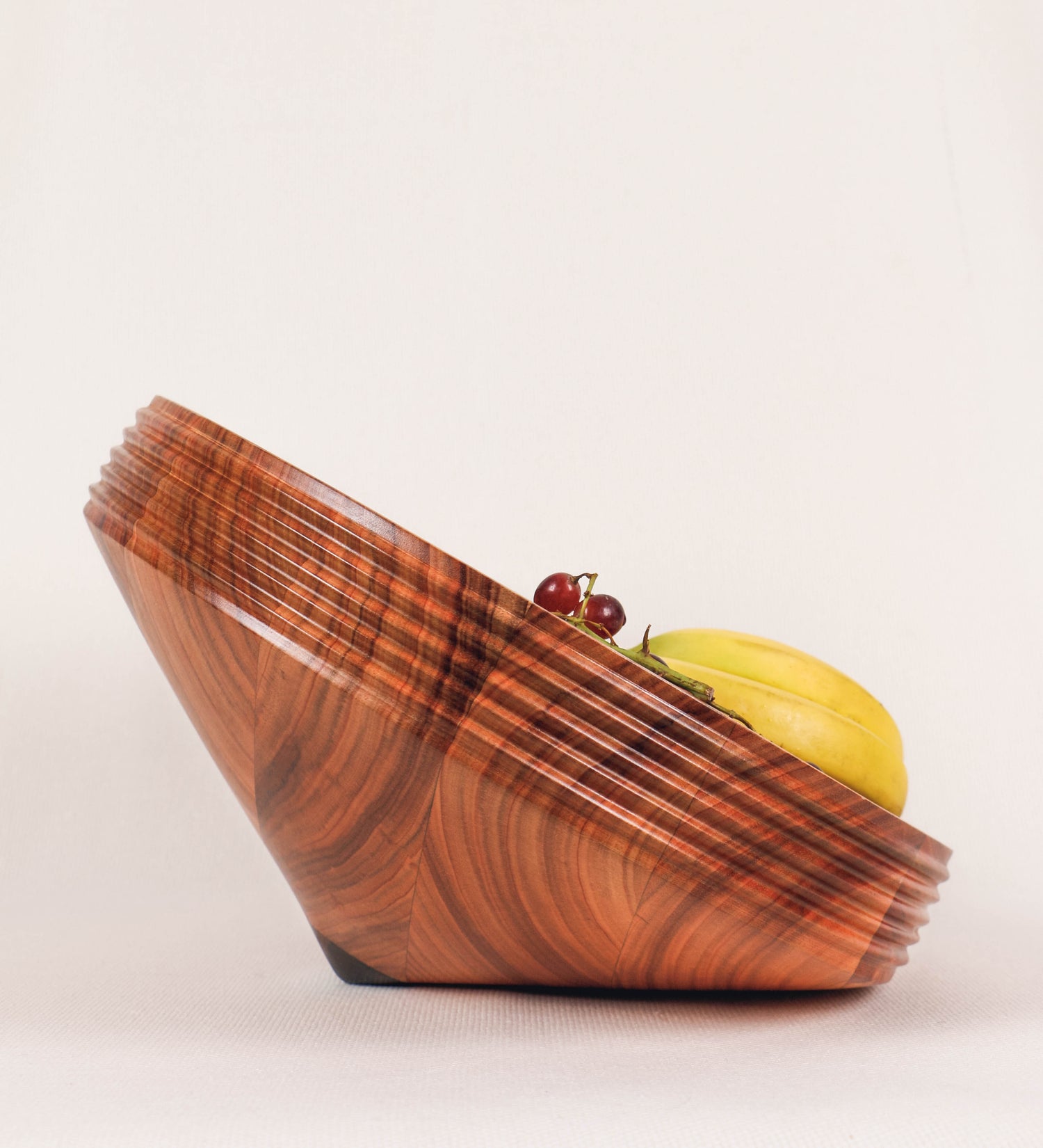 Fruteira com um design minimalista e distinto. A fruteira é de madeira de Acácia-Austrália, com uma tonalidade escura e verniz foodsafe semi-brilhante. A peça é de formato cónico e com um toque liso, semelhante a um pião, com uma ponteira em madeira também de tom mais escuro, quase negro. Apresenta ainda uma borda de cerca de 3 cm de altura, próxima da abertura da peça, decorada com relevos lineares dispostos horizontalmente, tipo ranhuras, que envolvem a fruteira.