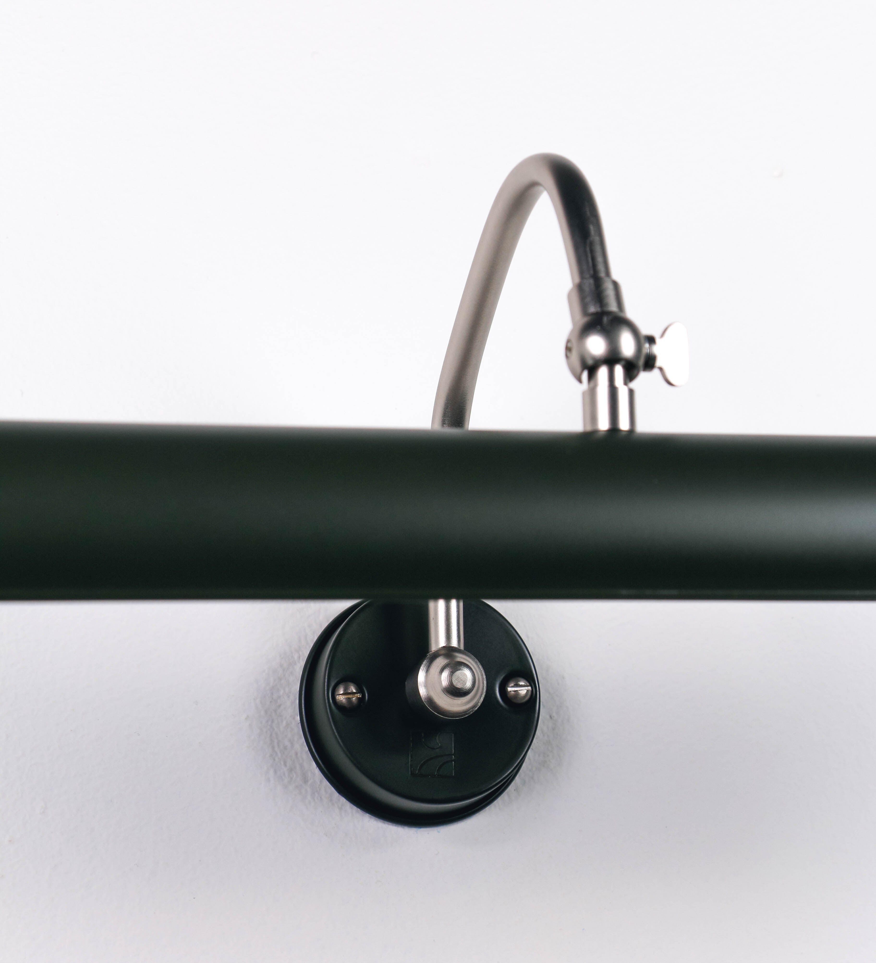 Aplique de parede que combina elementos clássicos com toques contemporâneos. O aplique é composto por uma base circular verde-azeitona, de onde se estende um braço curvo e gracioso, em níquel escovado, que suporta a barra principal. O braço contém um elemento, em níquel escovado, que permite apertar ou desapertar a barra para melhor manipular a direção da luz. A barra de 45cm de largura, é cilíndrica e alongada, verde-azeitona. As extremidades do cilindro são planas e do mesmo diâmetro.