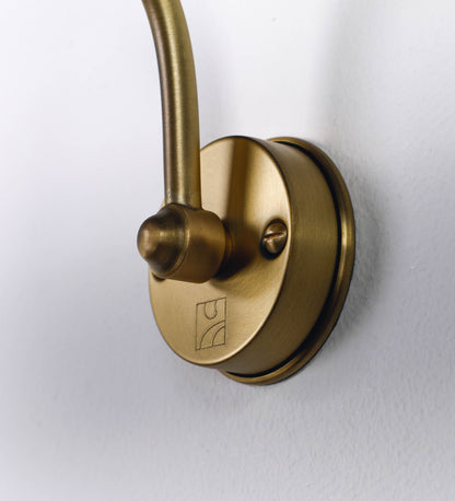 Aplique de parede que combina elementos clássicos com toques contemporâneos, todo em ouro velho. O aplique é composto por uma base circular de onde se estende um braço curvo e gracioso, que suporta a barra principal. O braço contém um elemento que permite apertar ou desapertar a barra para melhor manipular a direção da luz. A barra de 45cm de largura é cilíndrica e alongada. As extremidades do cilindro são planas e do mesmo diâmetro.