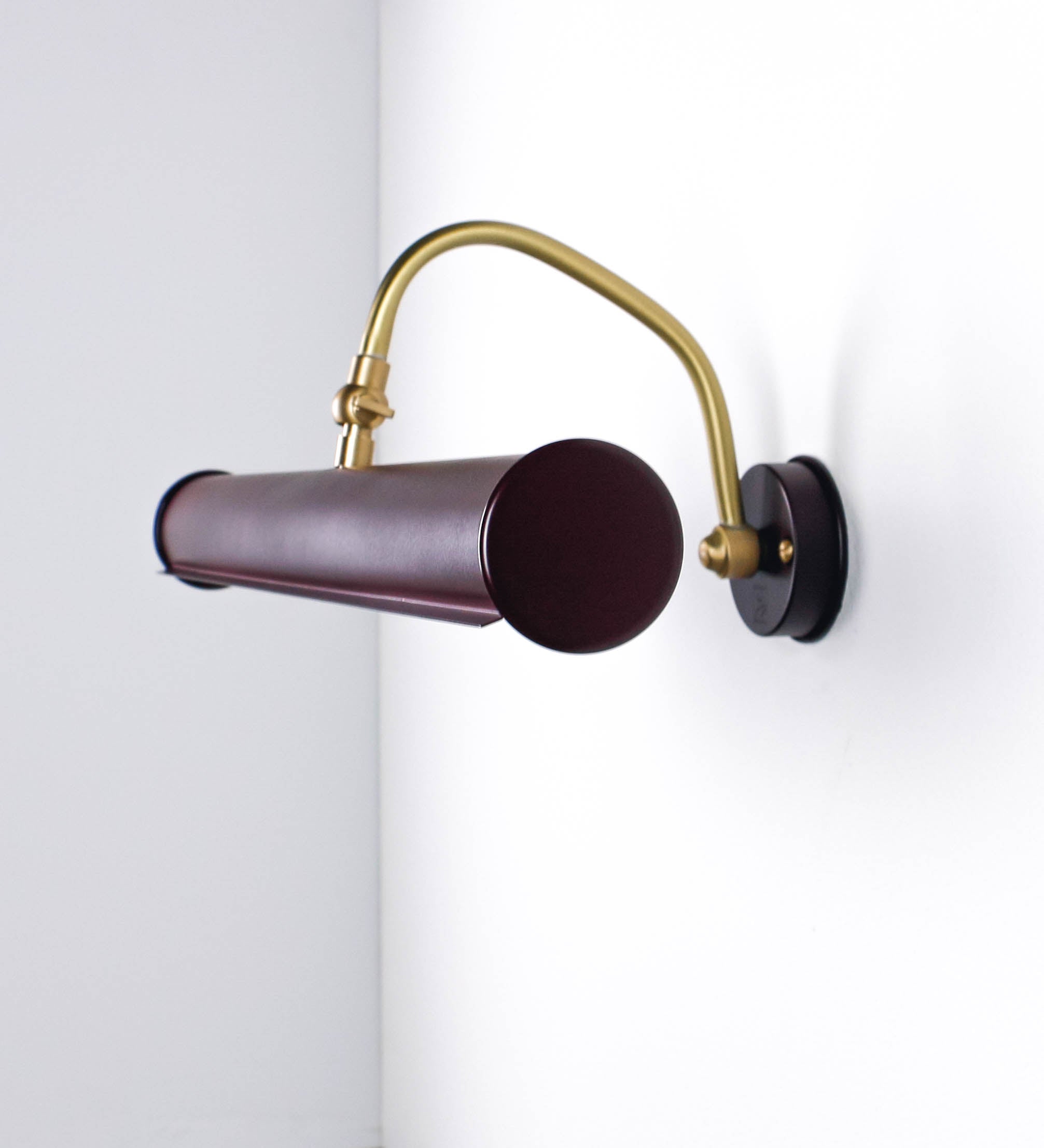 Aplique de parede que combina elementos clássicos com toques contemporâneos. O aplique é composto por uma base circular tom vinho, de onde se estende um braço curvo e gracioso, em latão escovado, que suporta a barra principal. O braço contém um elemento, em latão escovado, que permite apertar ou desapertar a barra para melhor manipular a direção da luz. A barra de 45cm de largura, é cilíndrica e alongada, tom vinho. As extremidades do cilindro são planas e do mesmo diâmetro.