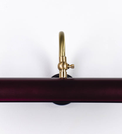 Aplique de parede que combina elementos clássicos com toques contemporâneos. O aplique é composto por uma base circular tom vinho, de onde se estende um braço curvo e gracioso, em latão escovado, que suporta a barra principal. O braço contém um elemento, em latão escovado, que permite apertar ou desapertar a barra para melhor manipular a direção da luz. A barra de 45cm de largura, é cilíndrica e alongada, tom vinho. As extremidades do cilindro são planas e do mesmo diâmetro.